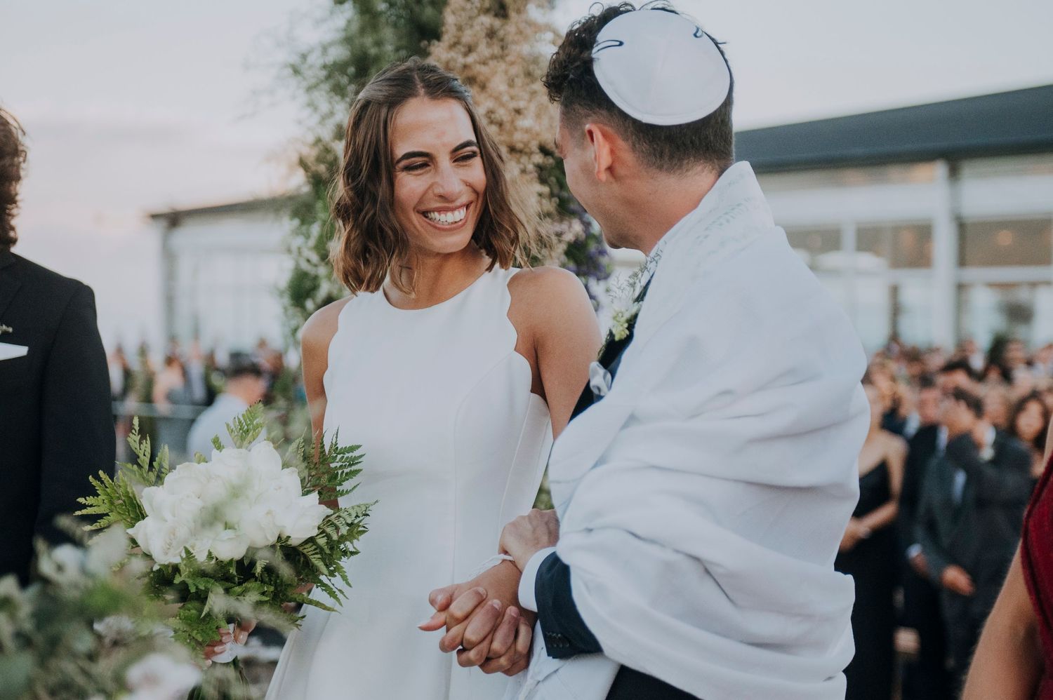 Smiling bride holding white bouquet exchanges vows with groom wearing kippah at outdoor Jewish wedding ceremony.