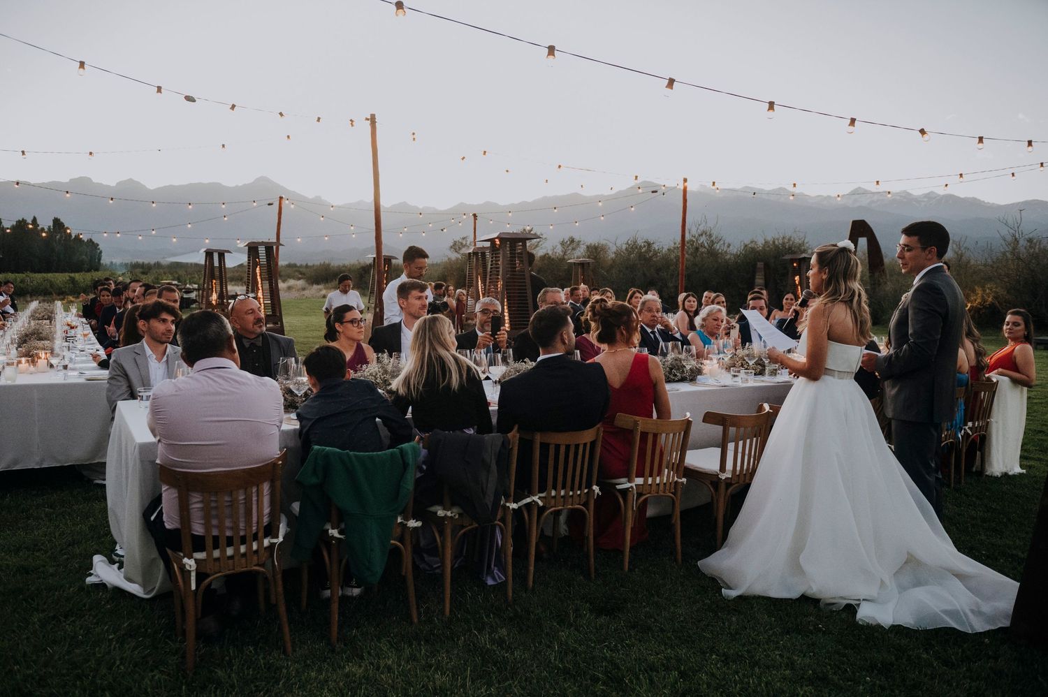 Bride and groom address seated guests at long outdoor reception table with mountain views.