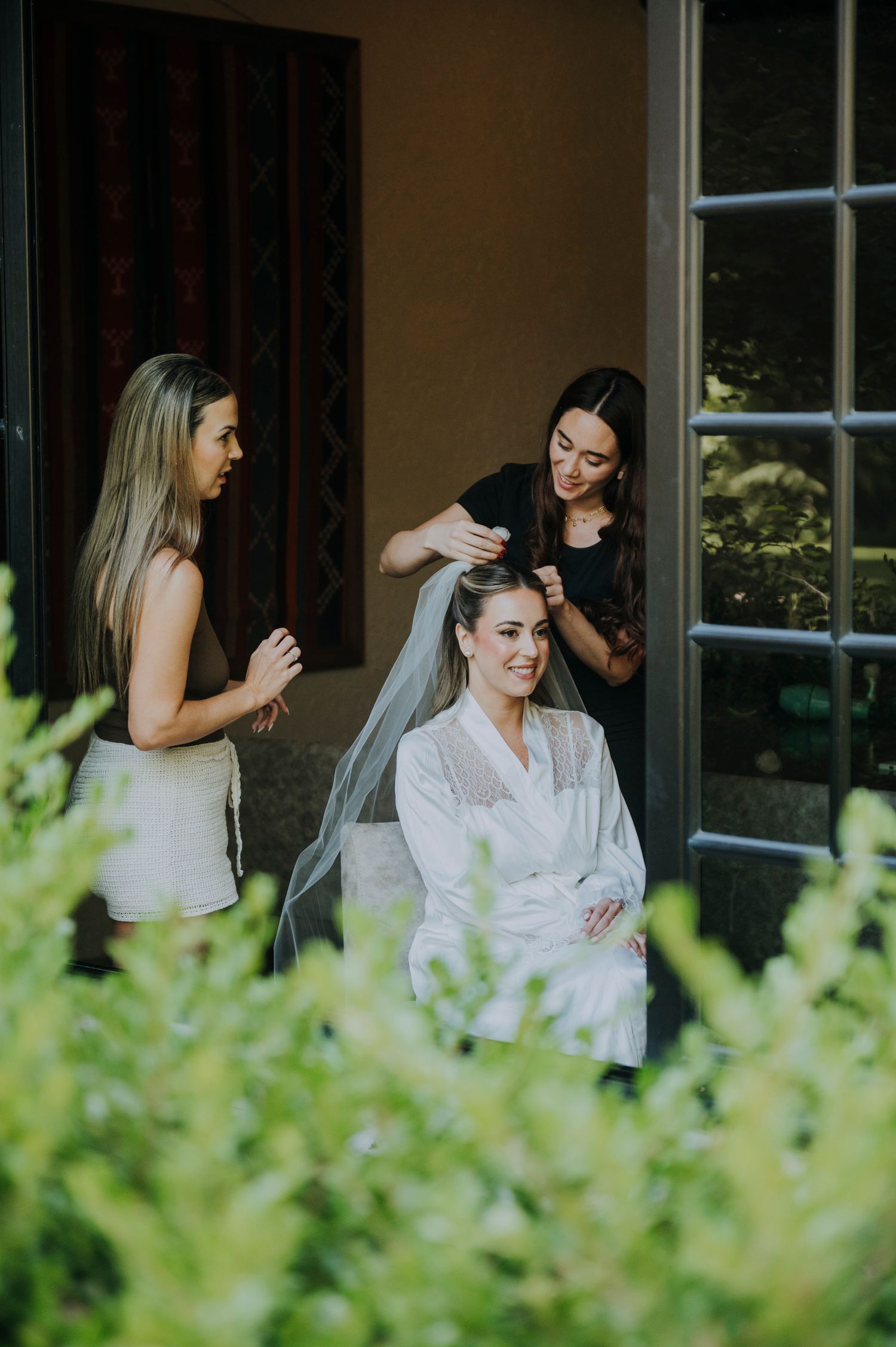 Bride in white robe smiles as stylist places veil while bridesmaid watches during wedding preparations.