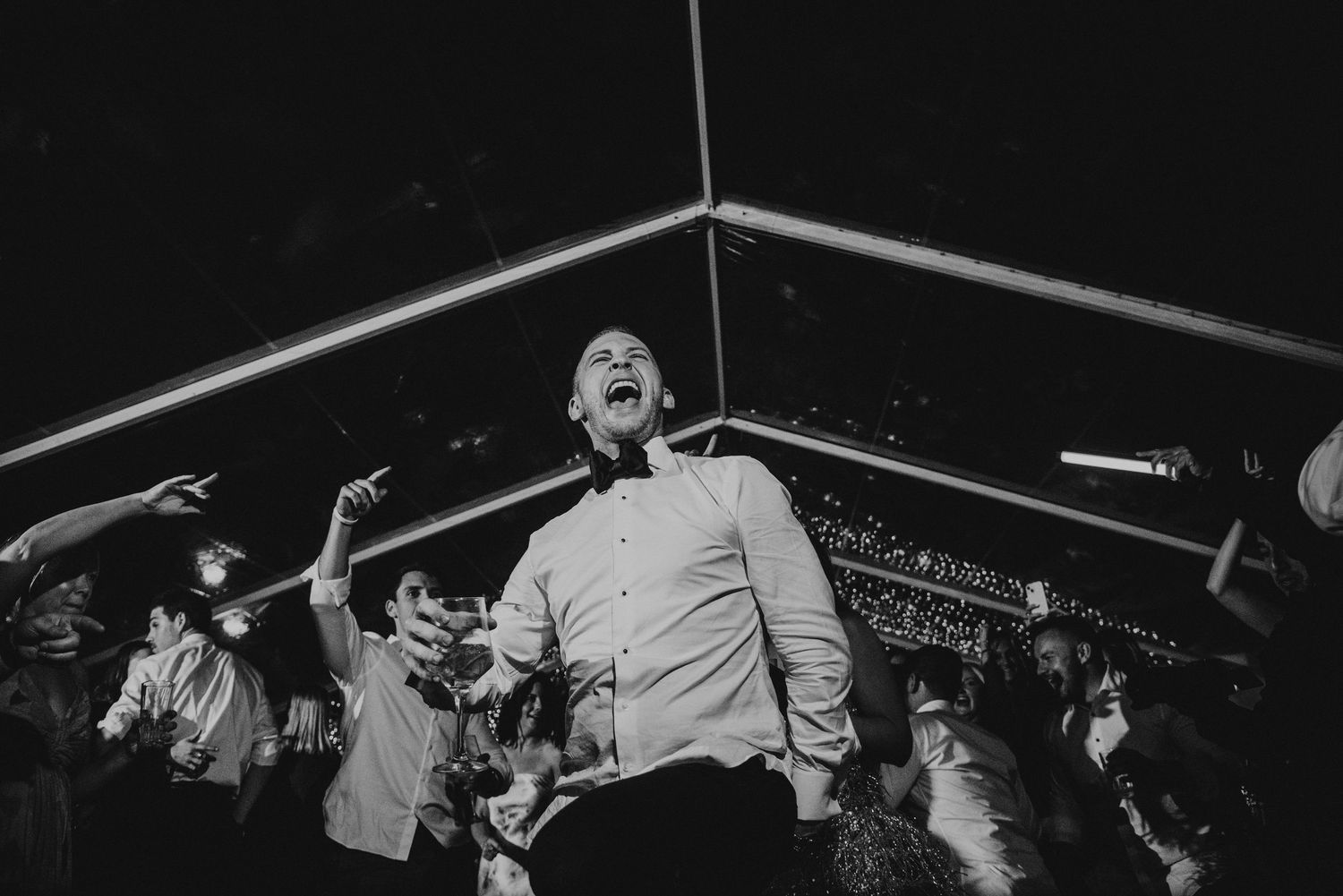 Black and white photo of energetic wedding guest dancing under string lights at reception.