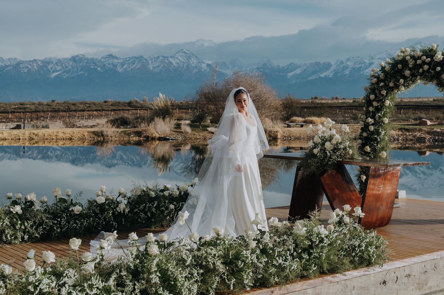 Bride in white gown and veil stands on floral-decorated platform with snow-capped mountains and lake backdrop.