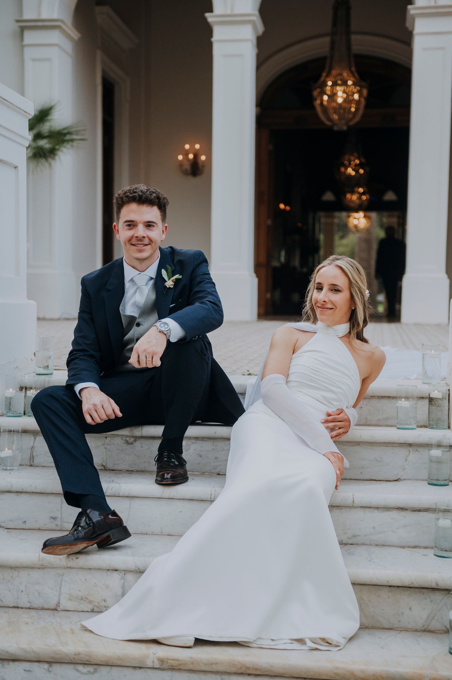 Bride and groom sitting on marble steps of elegant venue, smiling at camera.Couple portrait at sunset in Mendoza.