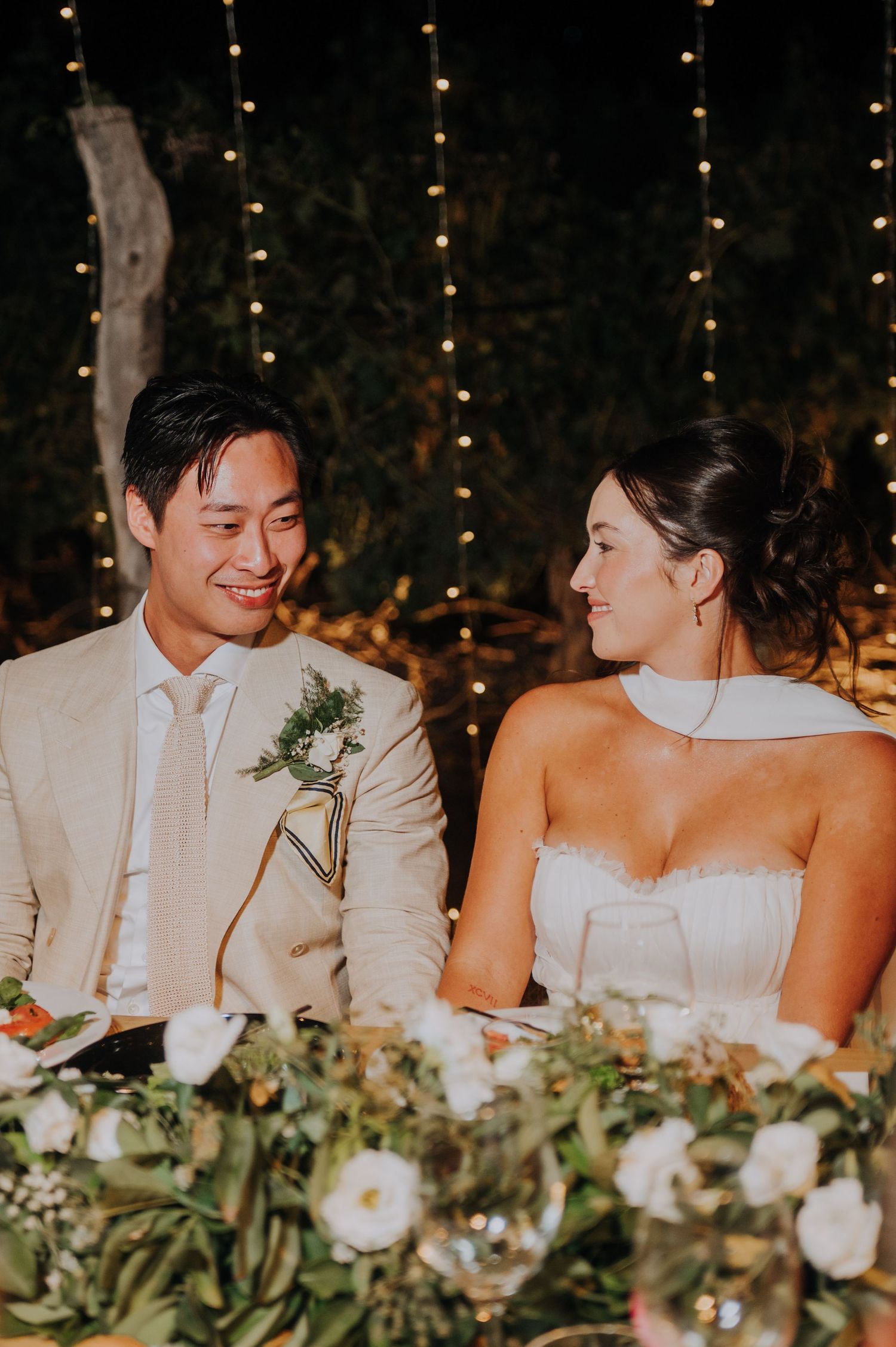 Bride and groom share a loving glance at sweetheart table with fairy lights and florals.