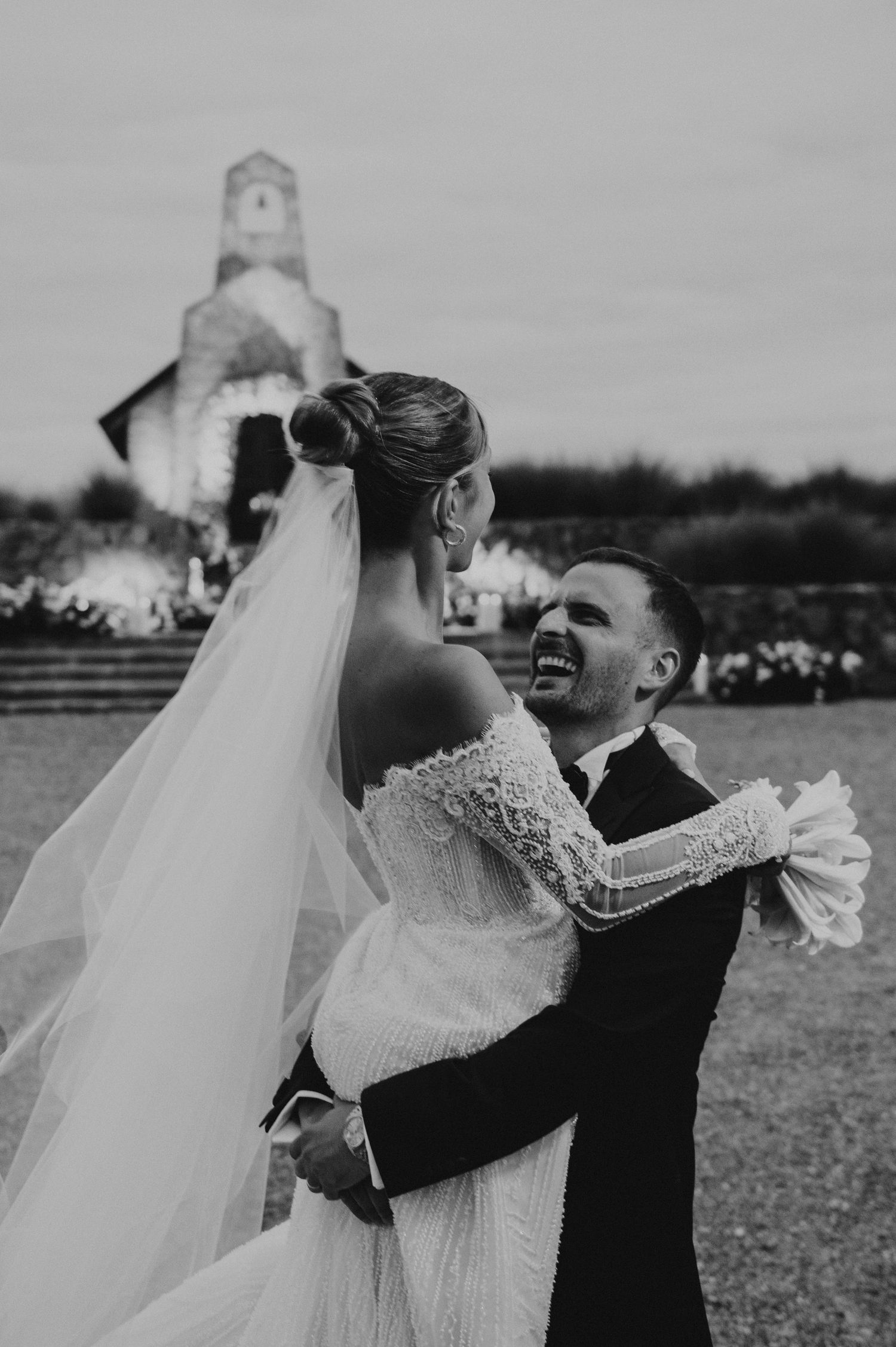 Black and white photo of groom embracing bride in lace gown with veil near a church after wedding ceremony.