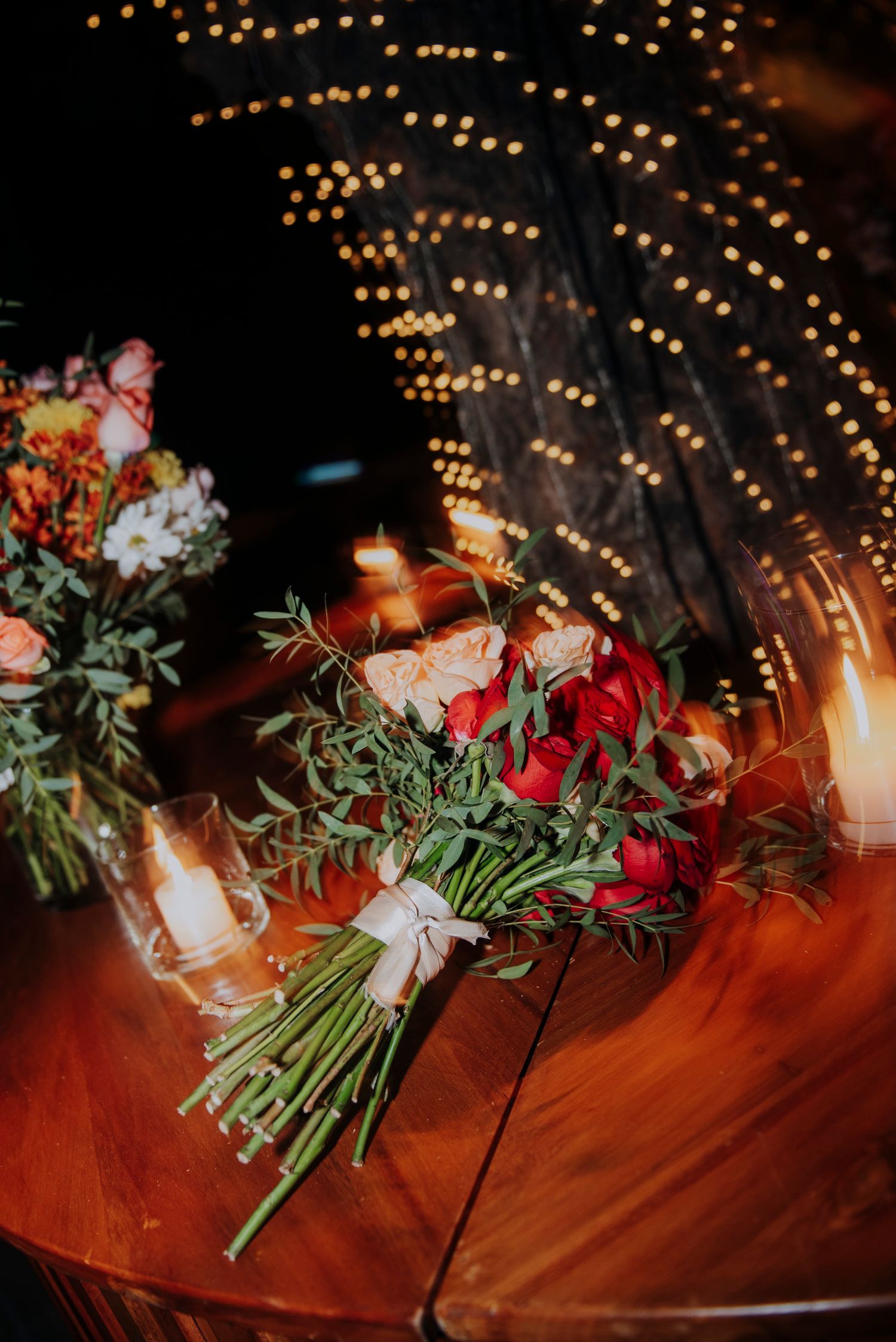 A romantic bridal bouquet of red roses and greenery on a wooden table lit by candles and fairy lights.