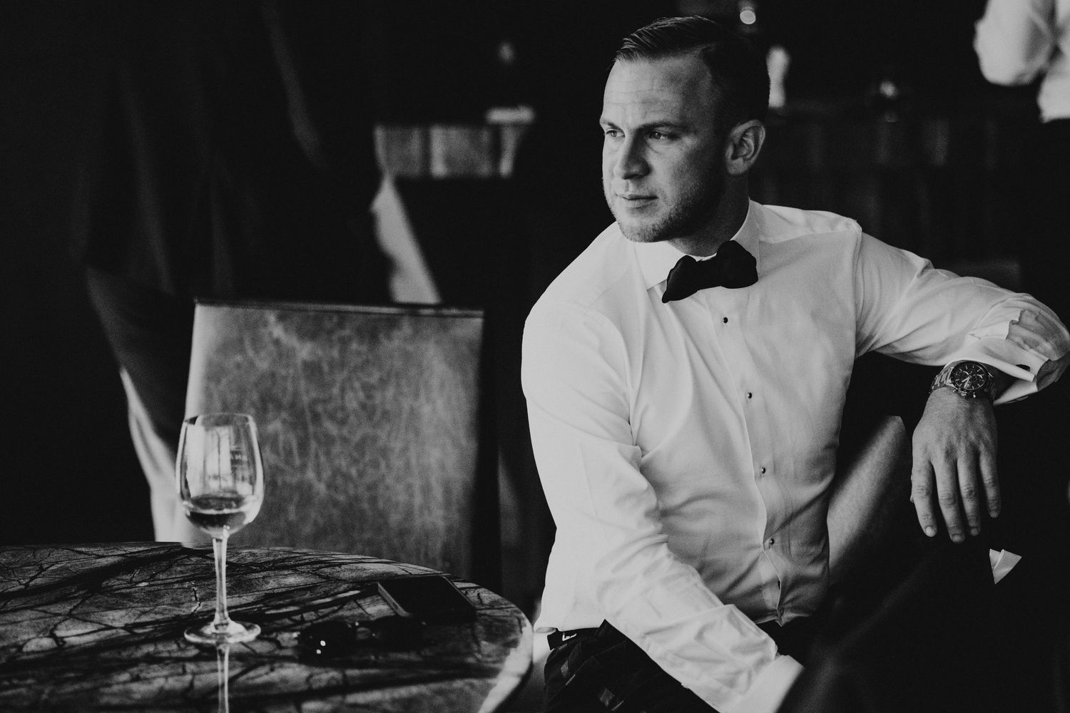 Elegant man in white shirt and bow tie sitting at a table with a wine glass in a dark upscale bar.