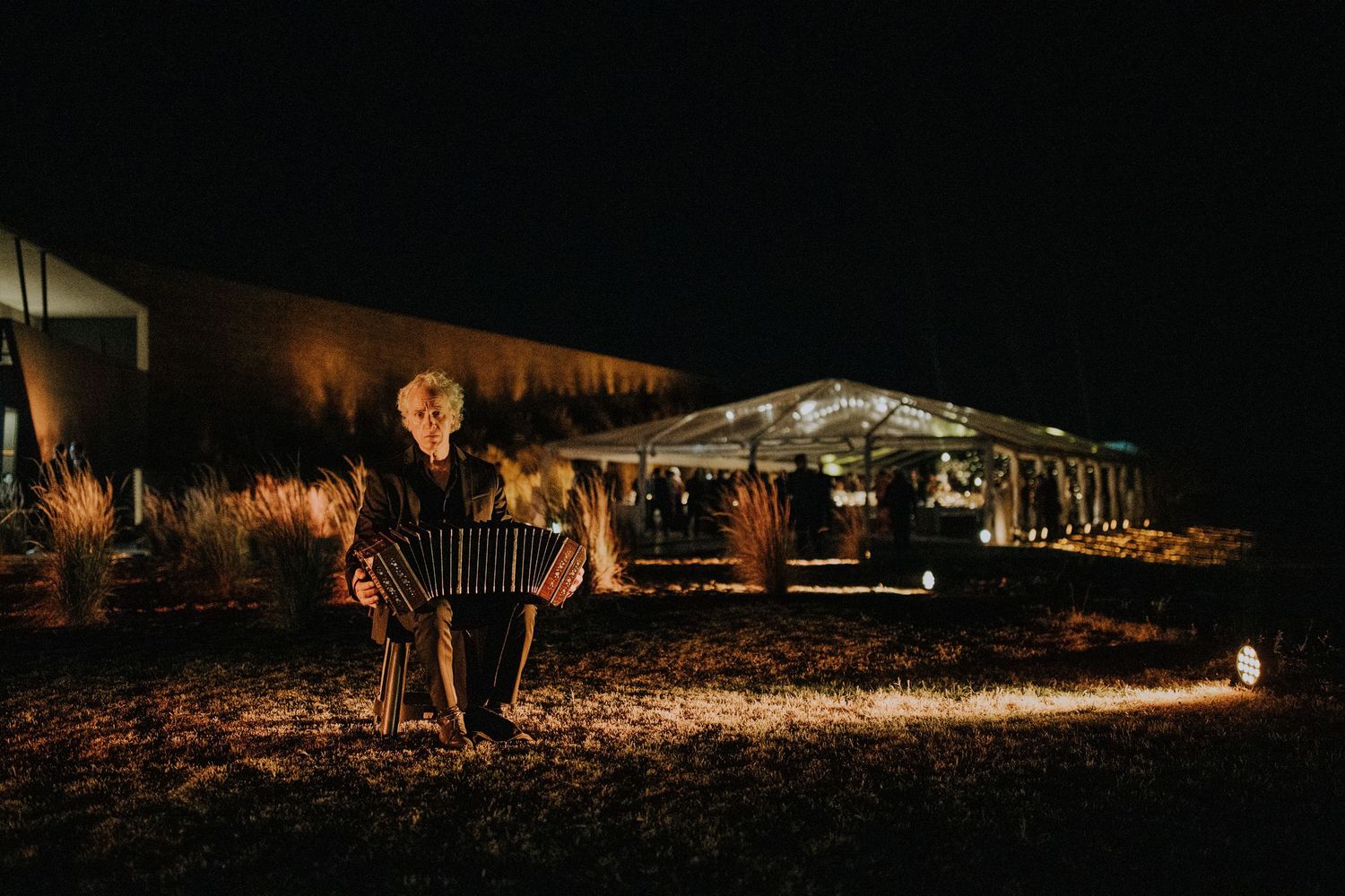 Musician playing accordion outdoors at night near an illuminated wedding reception tent.