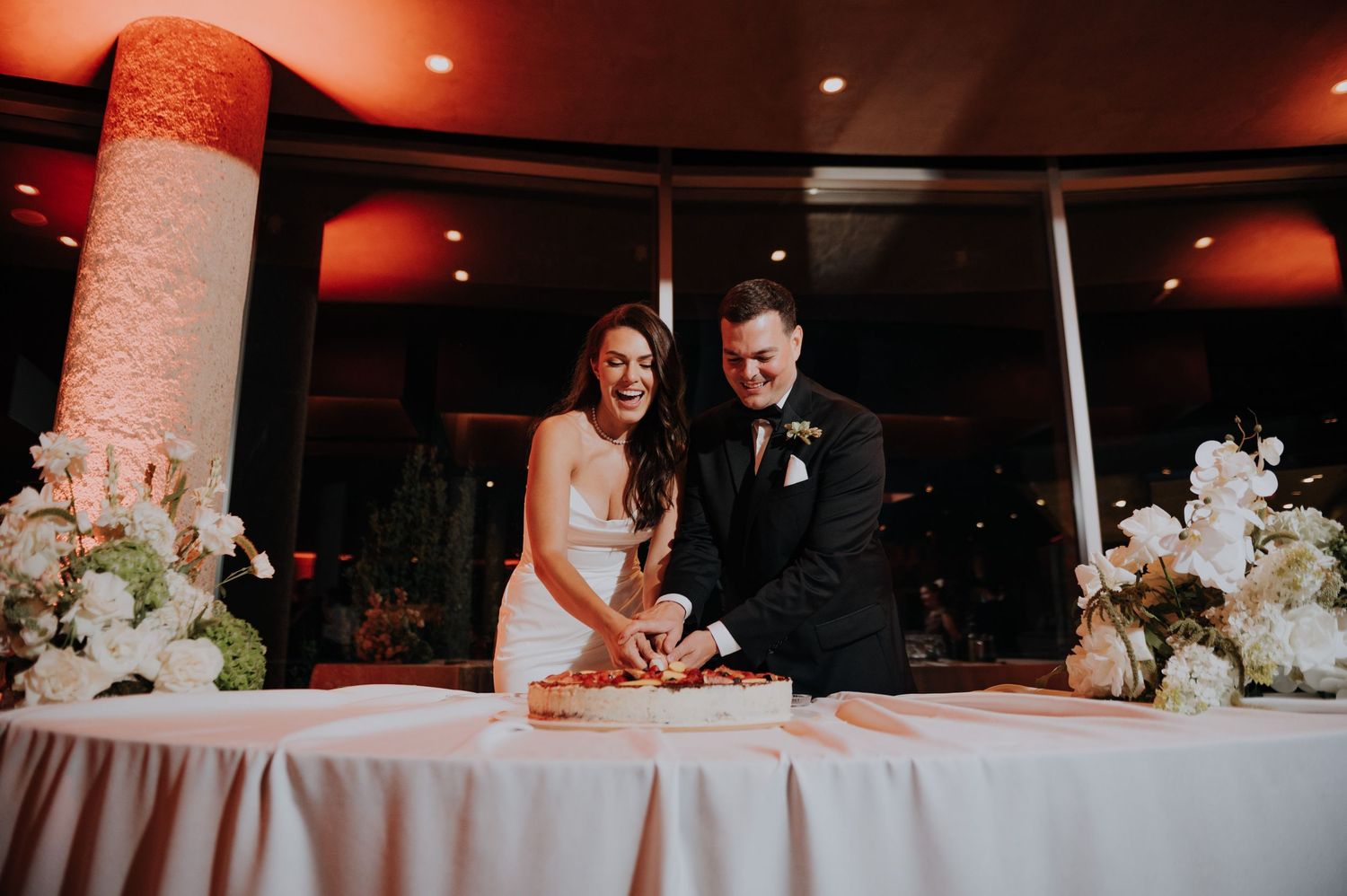Smiling bride and groom cut their elegant white wedding cake at reception venue.