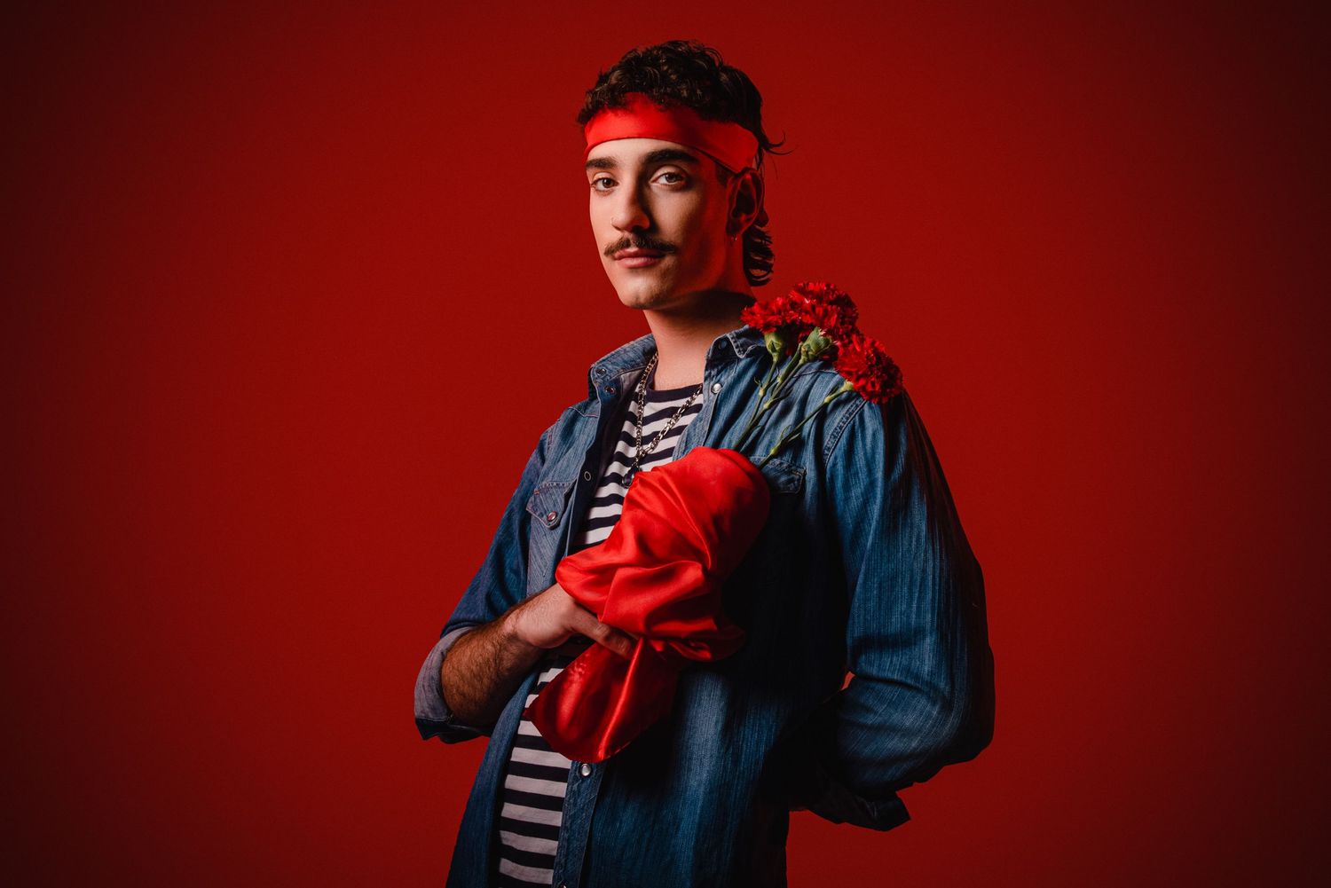 Santi Muk Young man in denim jacket holding red roses and fabric against a bold red background.