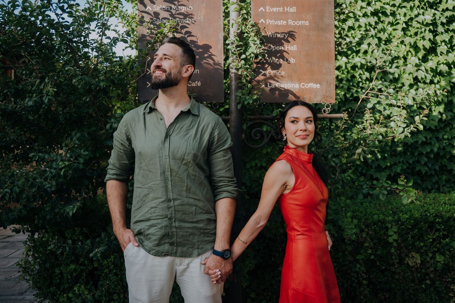 Couple holding hands outdoors, man in green shirt and woman in red dress, posing near ivy-covered wall.