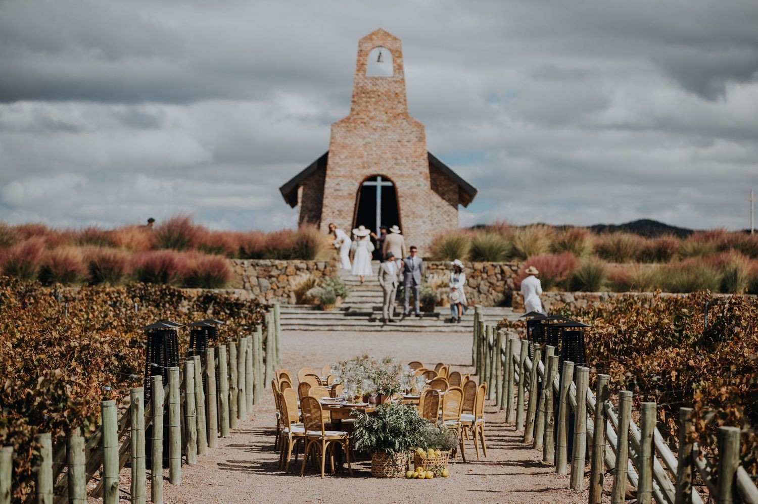 Rustic vineyard wedding ceremony with stone chapel, rattan chairs, and floral table setting under dramatic cloudy skies.