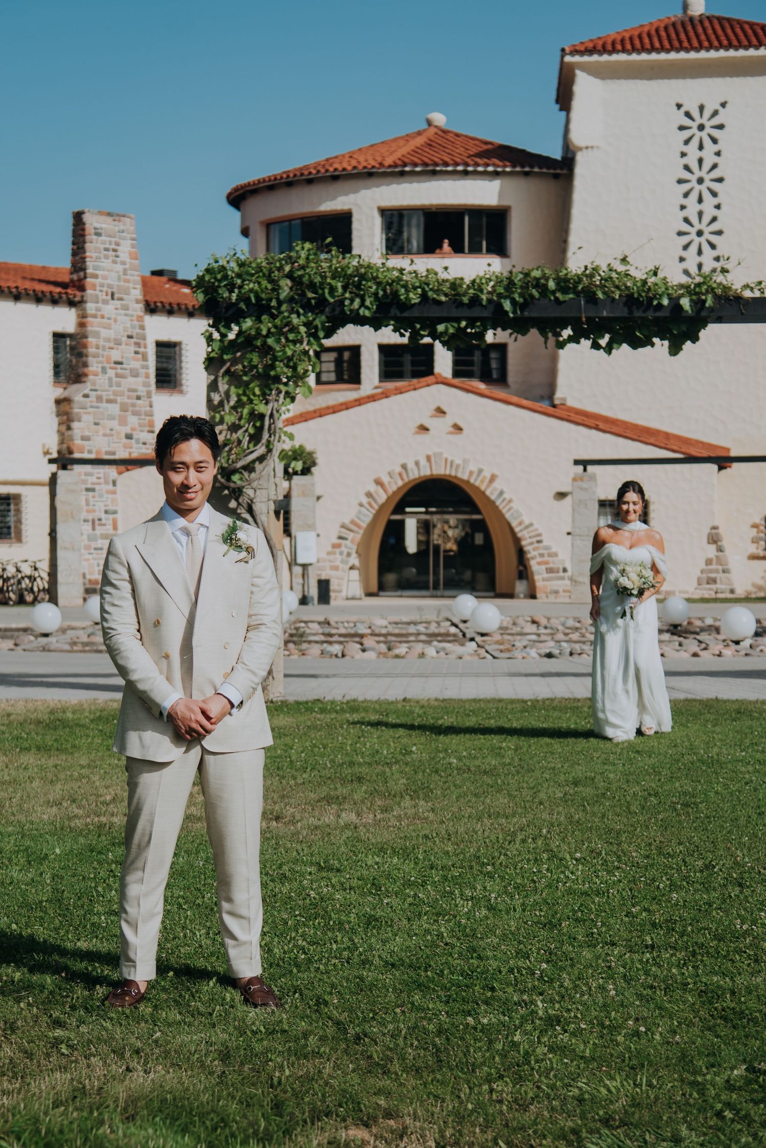 Groom in beige suit stands on lawn before Spanish-style wedding venue as bride approaches in background.