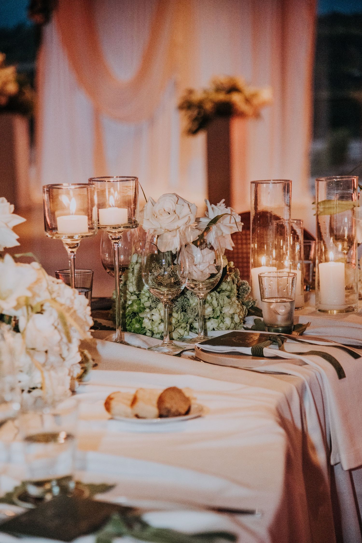 Elegant wedding reception table with white floral centerpieces, candles, and blush drapery.