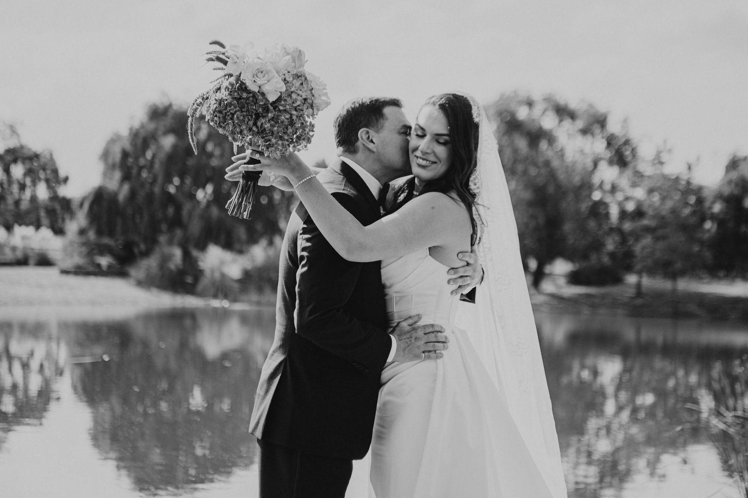 Black and white photo of groom kissing bride's cheek by a pond, bride smiling holding floral bouquet.