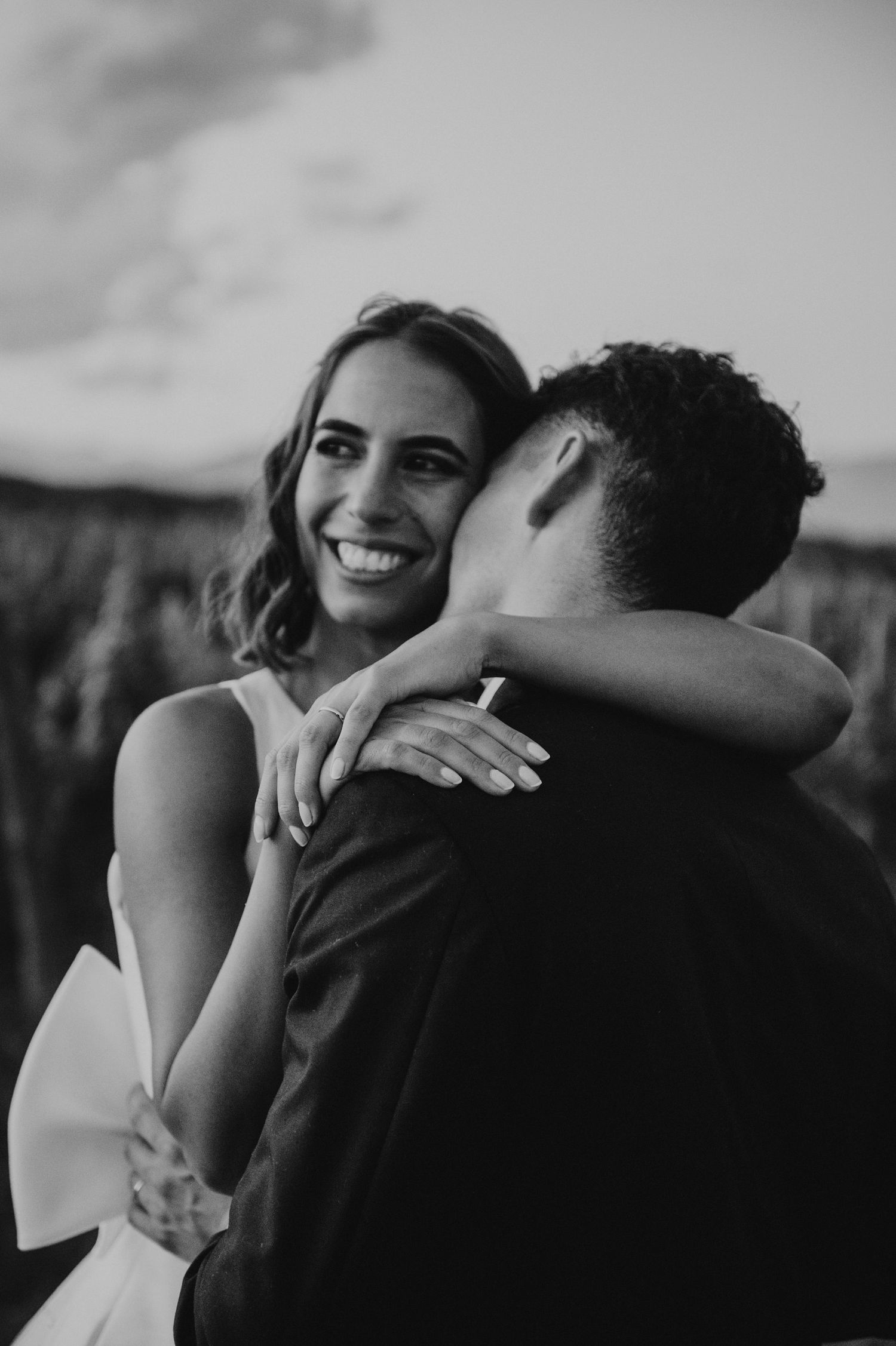 Black and white photo of smiling bride embracing groom outdoors at golden hour.