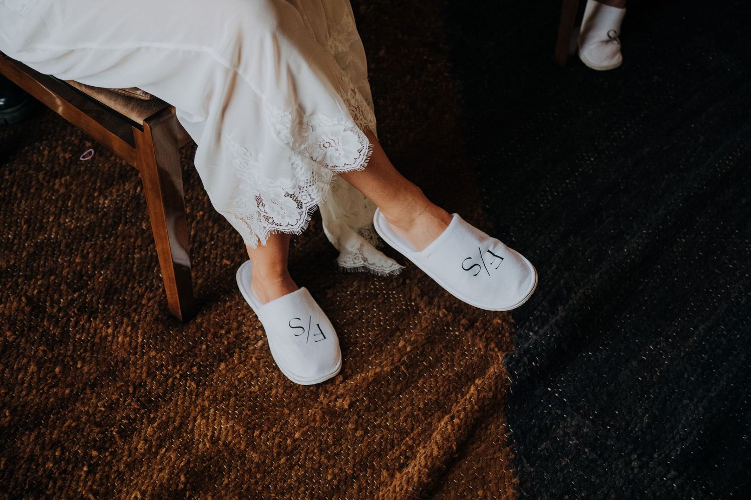 Bride wearing white monogrammed SA slippers with lace wedding dress hem visible while seated on wooden chair.