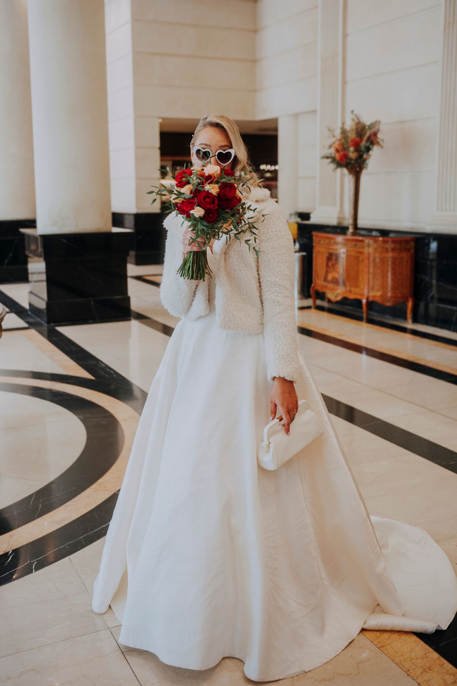Blonde bride in white gown and jacket holds red bouquet wearing heart-shaped sunglasses indoors.