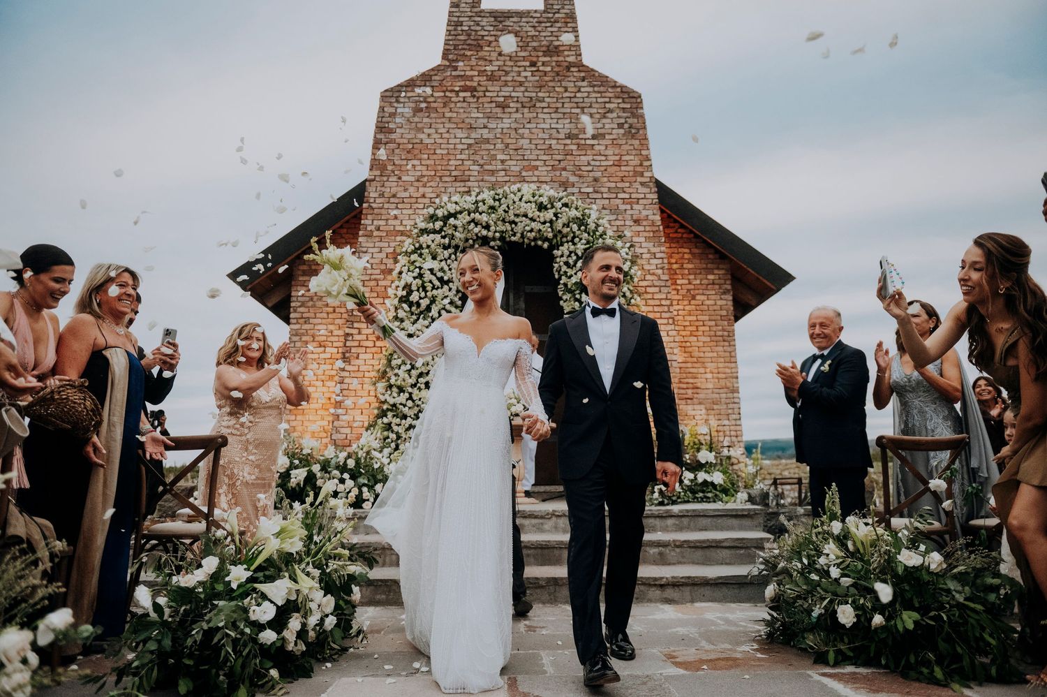 Bride and groom walk down the aisle as guests toss flower petals outside a rustic brick chapel wedding venue.