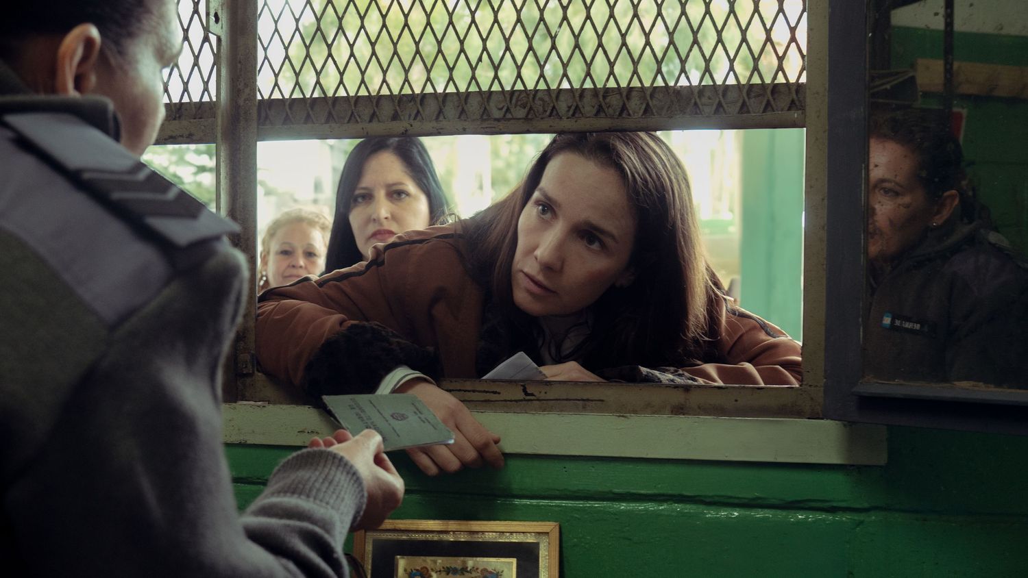 Natalia OreiroYoung woman leaning through a green ticket window handing over money in a film scene.