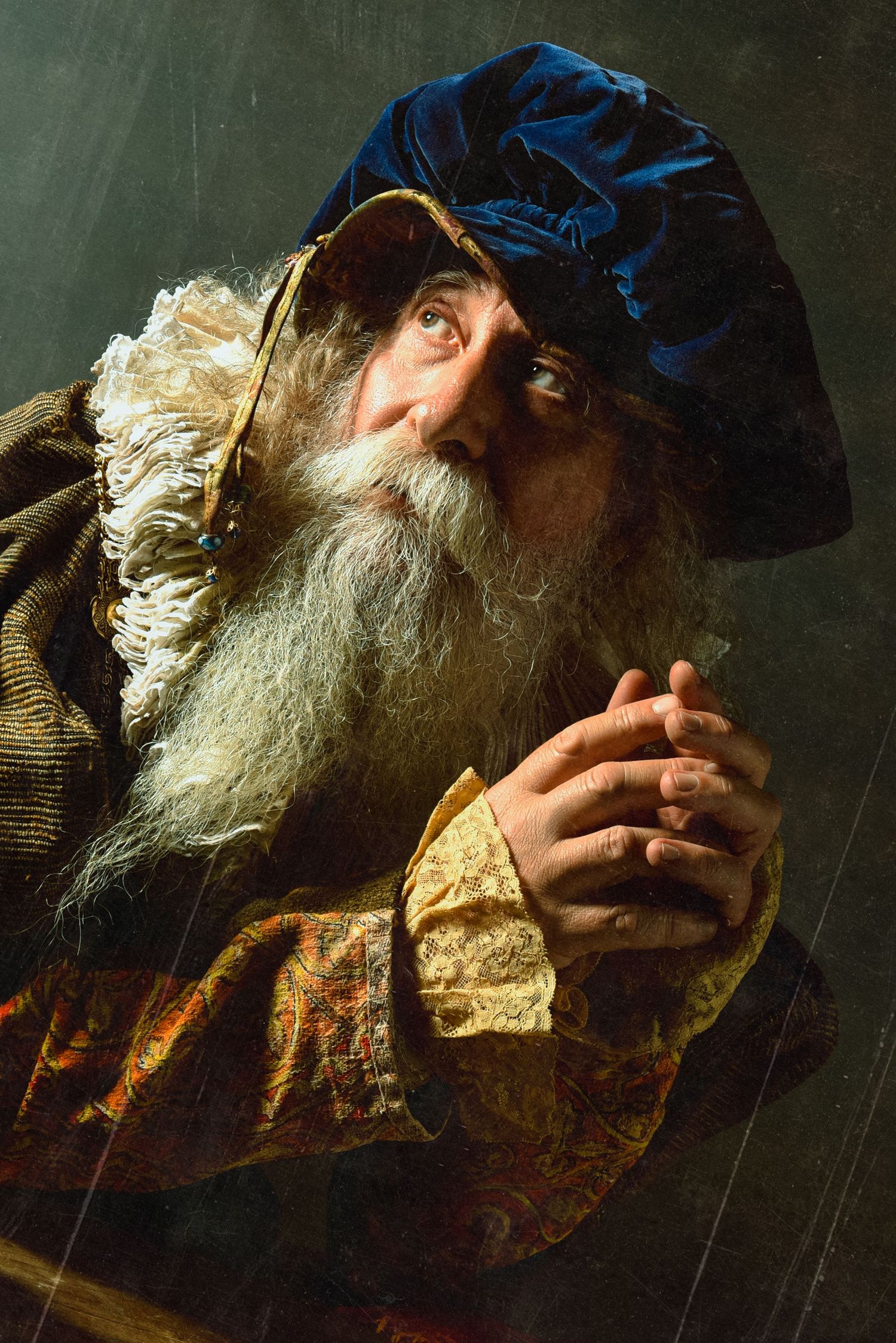 Elderly man with long white beard in vintage costume with blue velvet hat clasping hands dramatically