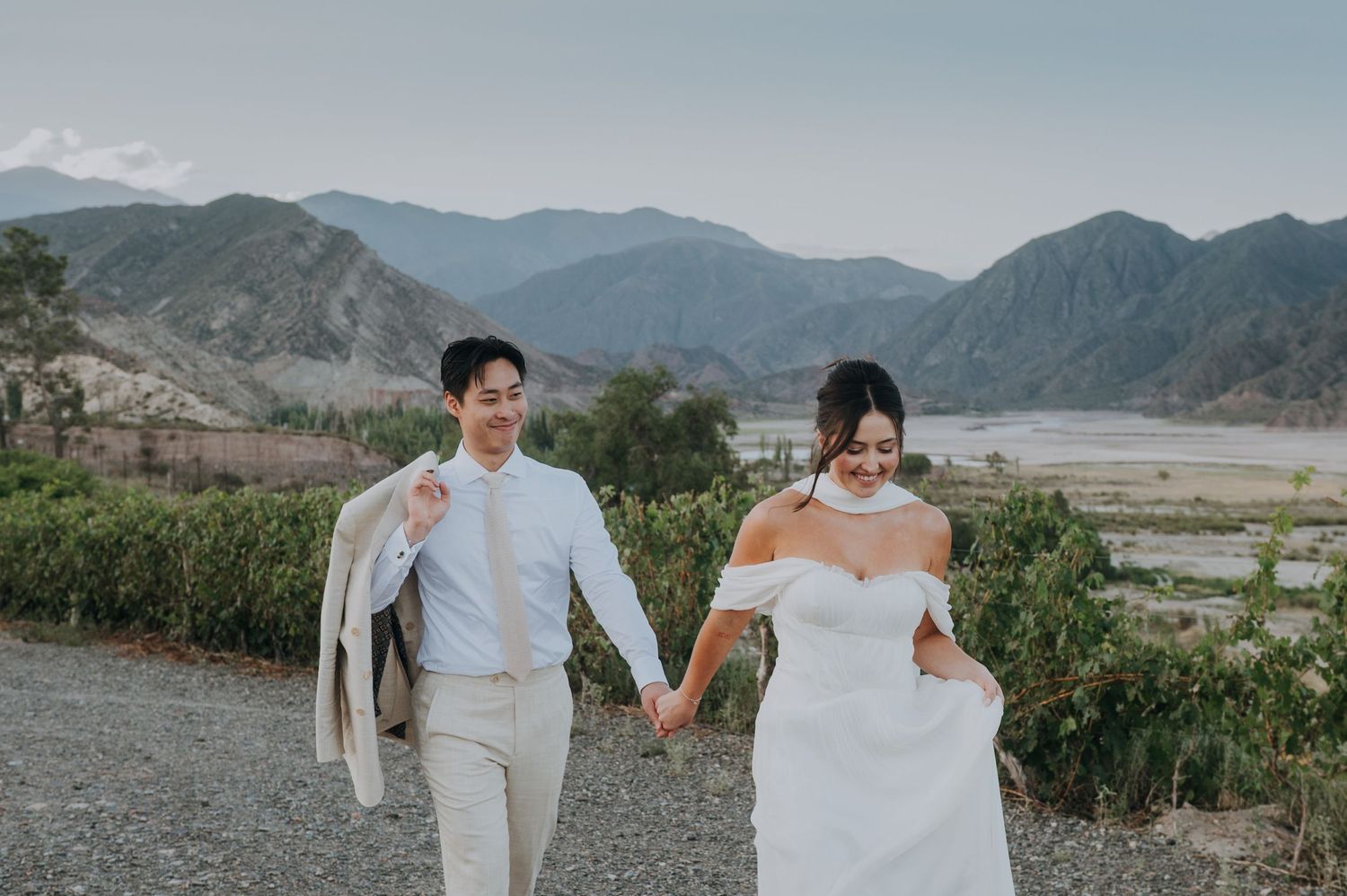 Happy bride and groom walking hand in hand through a scenic mountainous vineyard landscape on their wedding day.