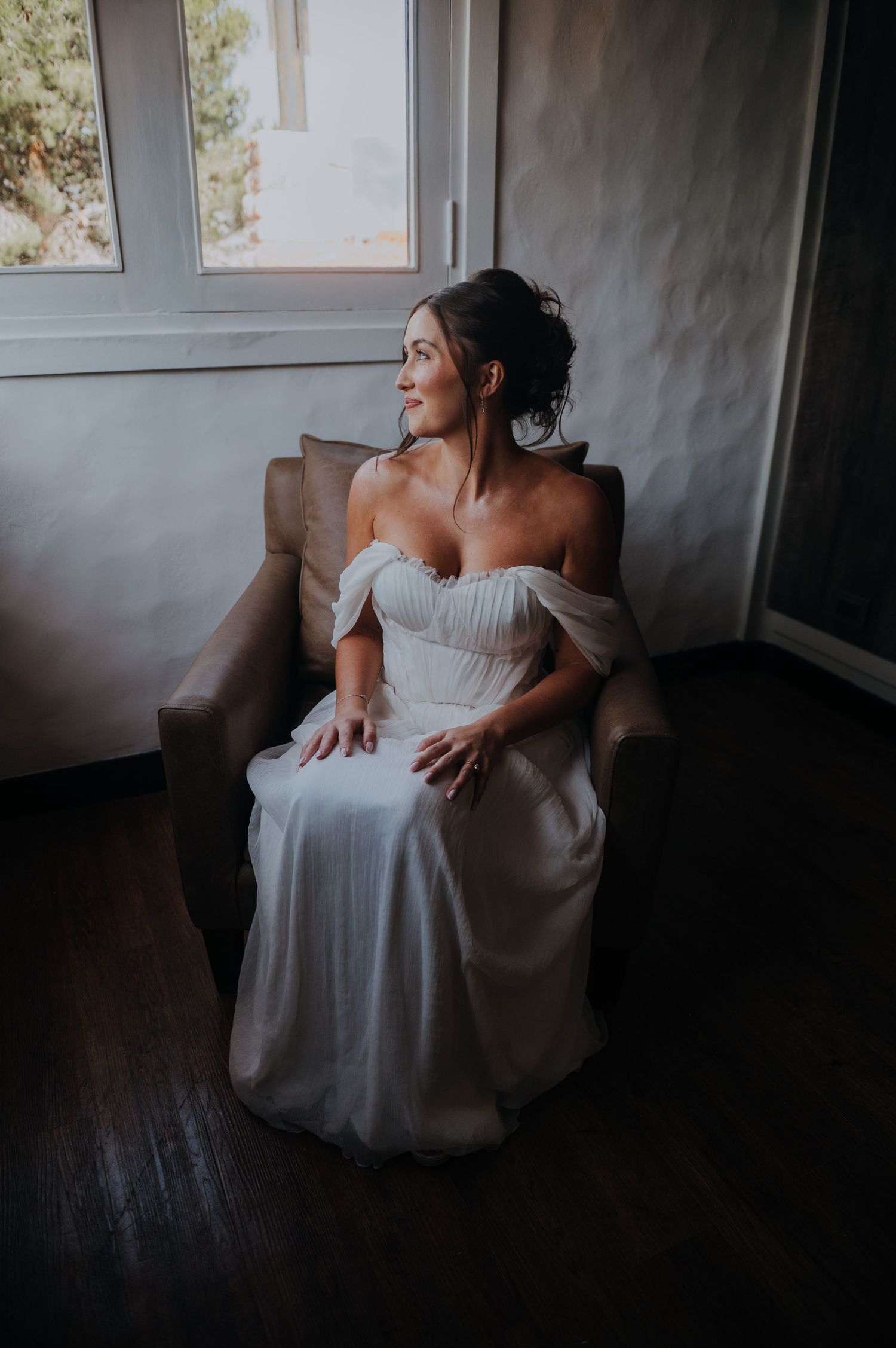 Radiant bride wearing an off-shoulder white wedding dress seated in an armchair smiling while looking out a sunlit window.