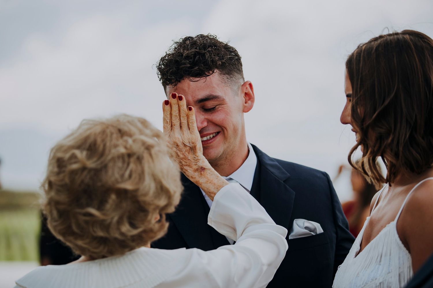 Emotional groom smiling as older woman touches his face tenderly at wedding ceremony.