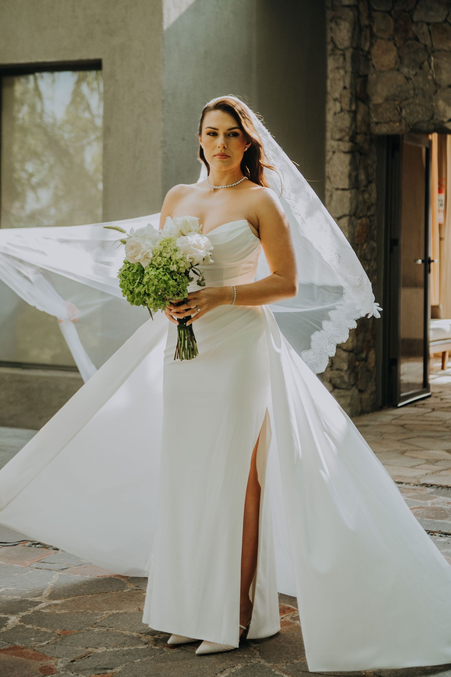 Elegant bride in strapless white gown with slit holds green and white bouquet as veil flows in the breeze.