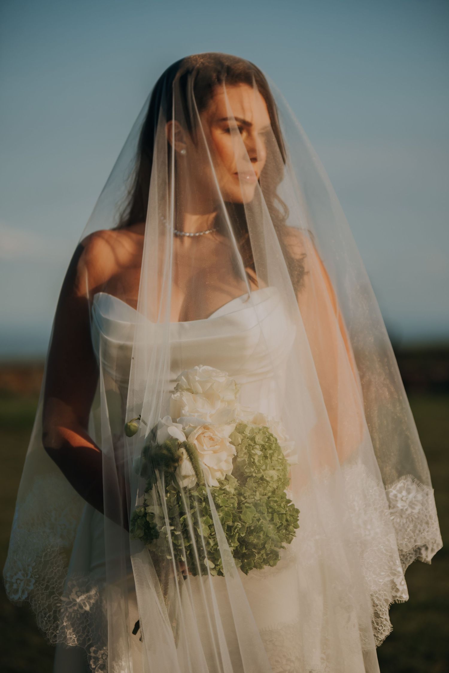 Elegant bride wearing lace-trimmed veil holding white and green hydrangea bouquet at sunset.