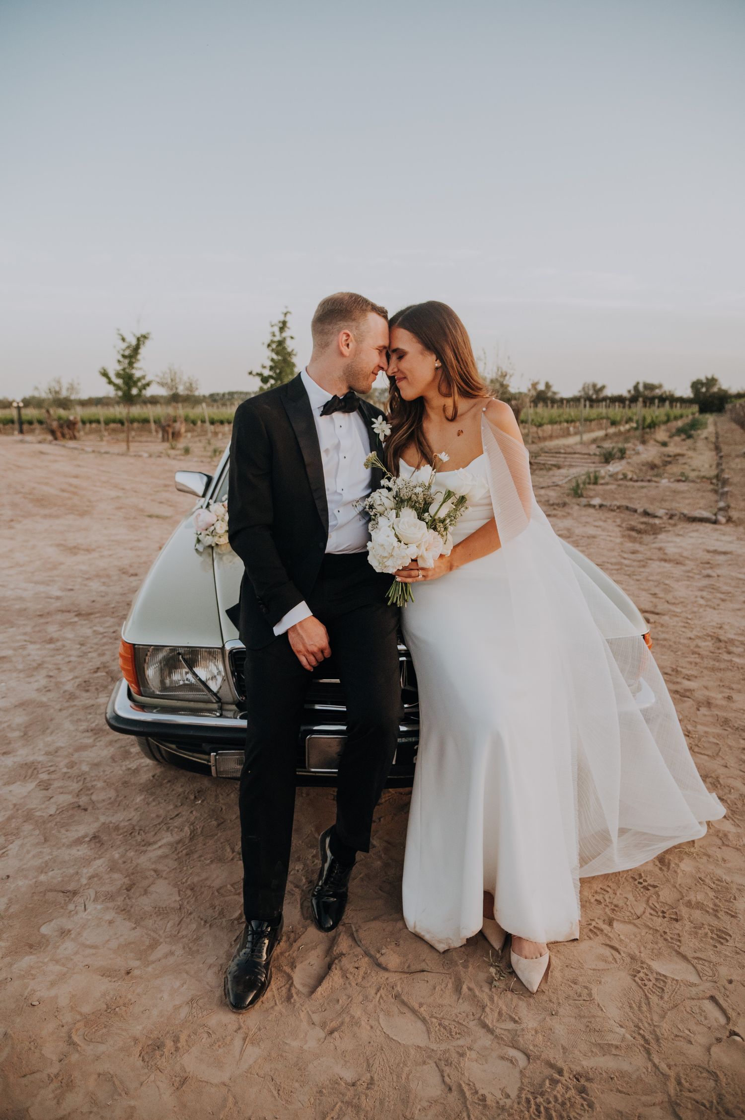 Bride and groom lean against a vintage car in a vineyard, sharing an intimate moment in wedding attire.