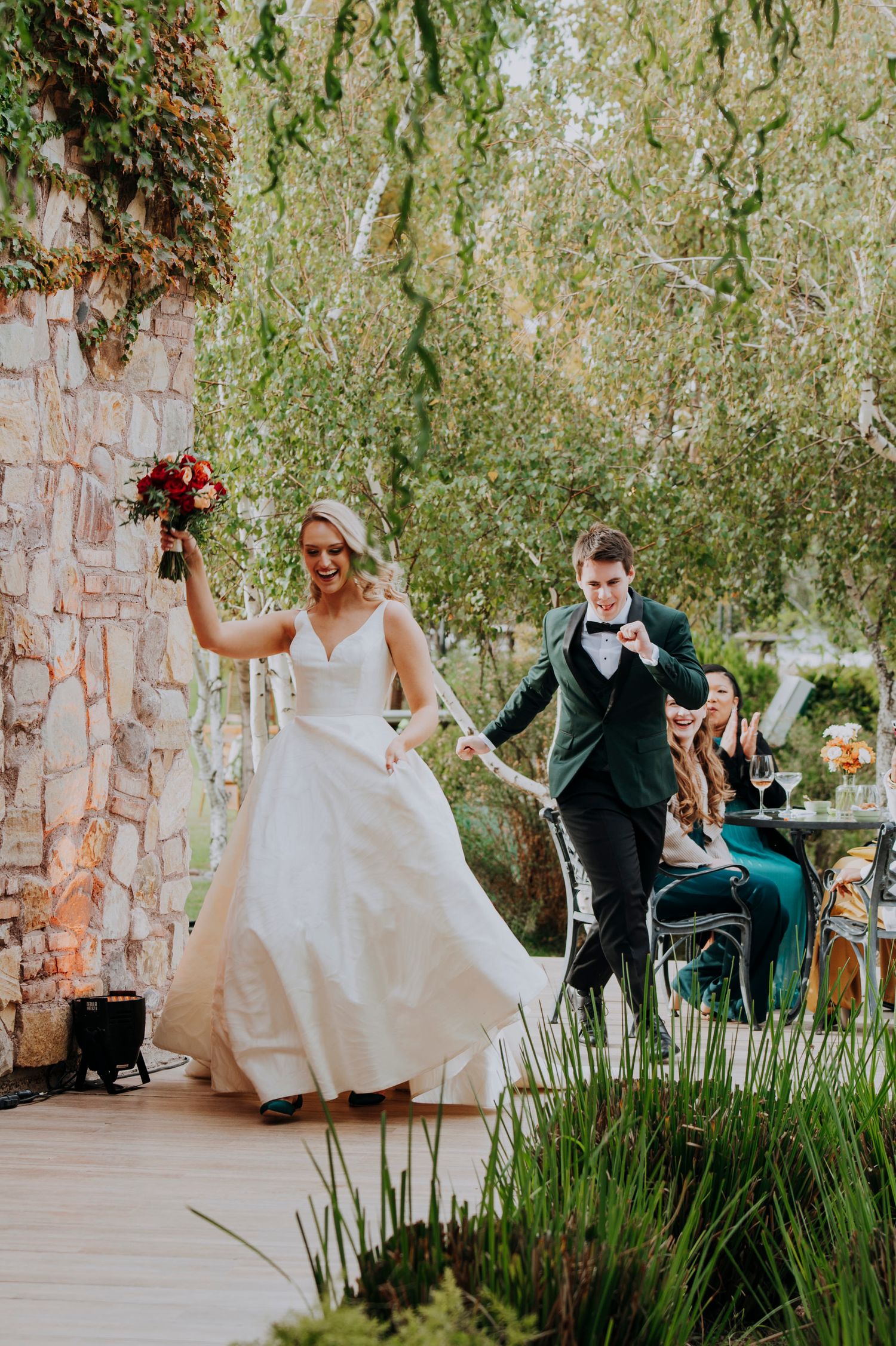 Joyful bride in white gown and groom in green suit making grand entrance holding red bouquet aloft.