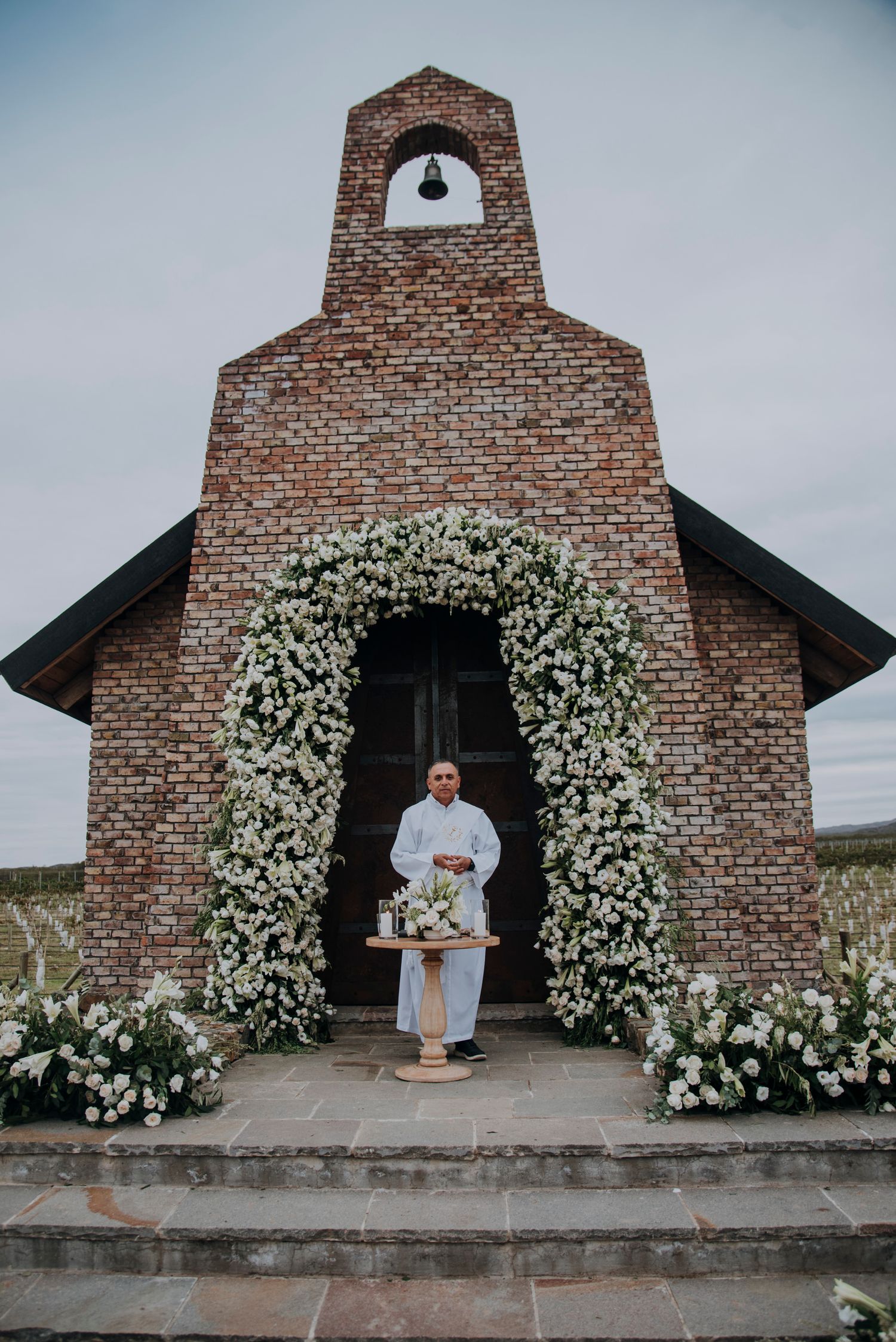 Officiant stands beneath a white floral arch at a brick chapel entrance, surrounded by lush white flower arrangements.