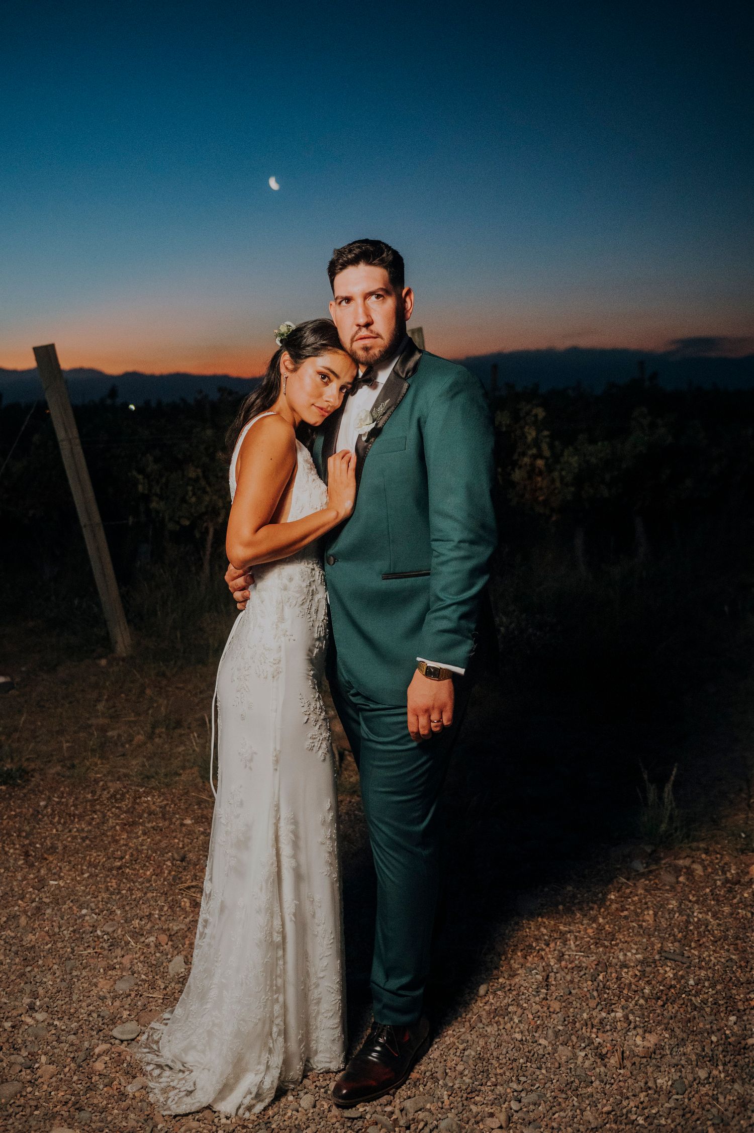 Wedding couple embracing at dusk in vineyard mendoza, groom in teal suit, bride in lace dress with crescent moon.w