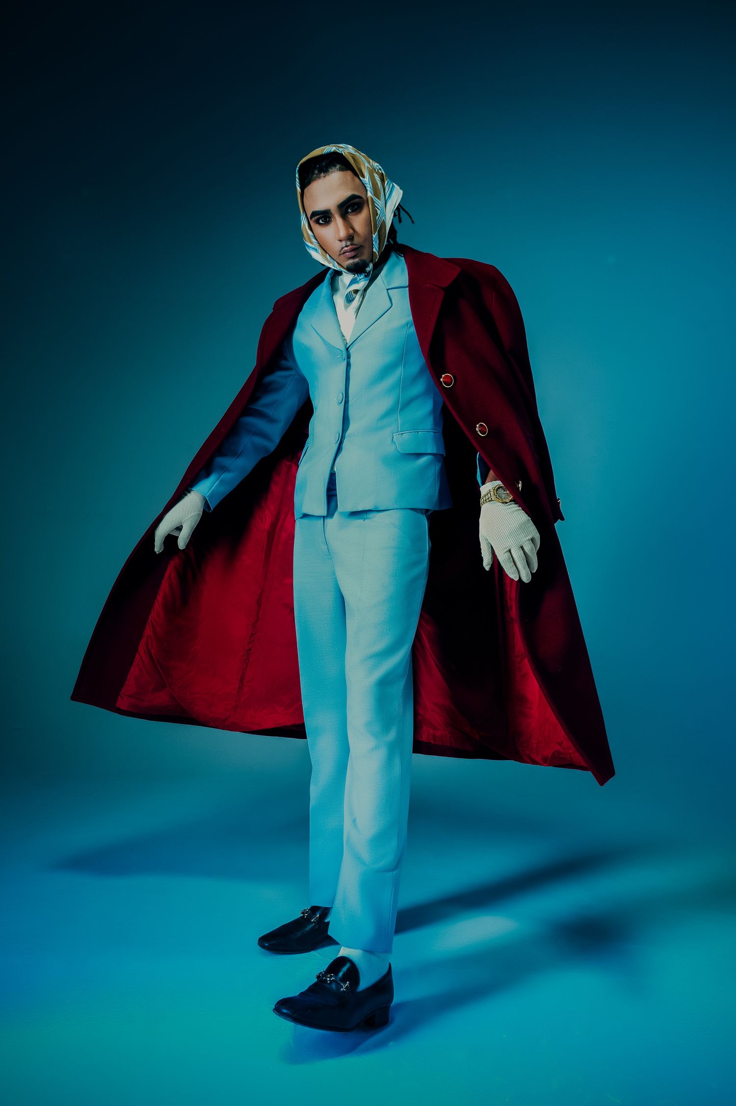 Man in light blue suit and red cape poses dramatically against a dark blue studio background.