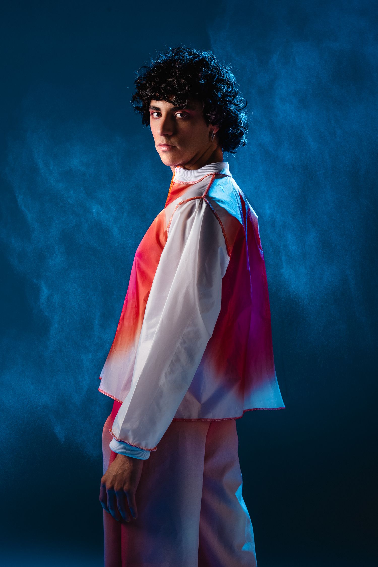 Young man with curly hair in colorful windbreaker posing against smoky blue studio background.