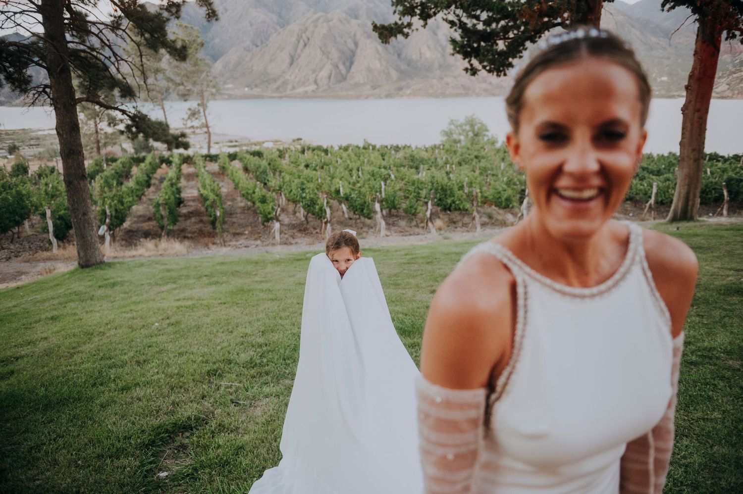Bride smiling at camera while groom hides under veil in vineyard setting. Wedding reception at a Mendoza winery.