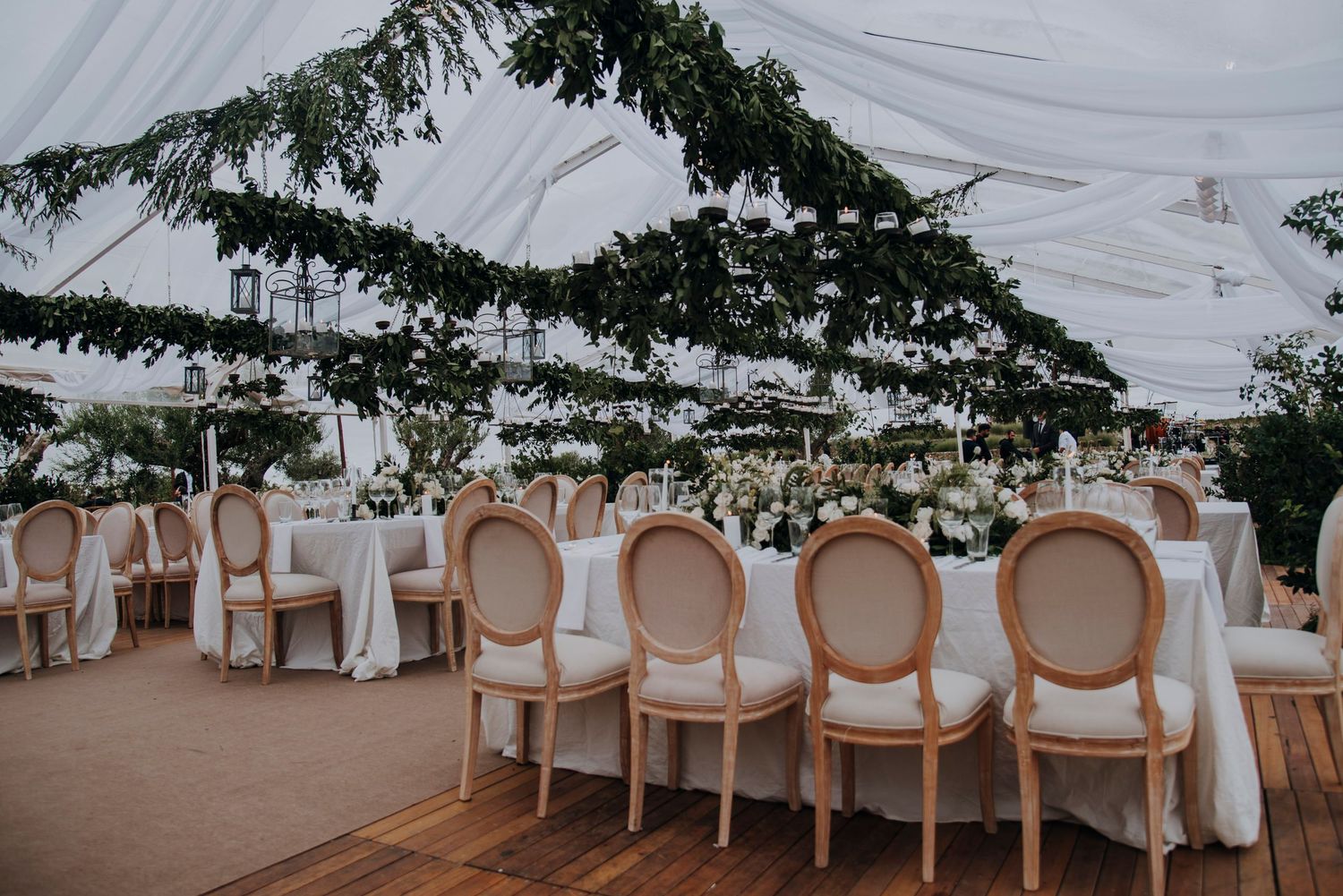 Elegant outdoor wedding reception under a white draped tent with greenery, round-back chairs, and white floral centerpieces.