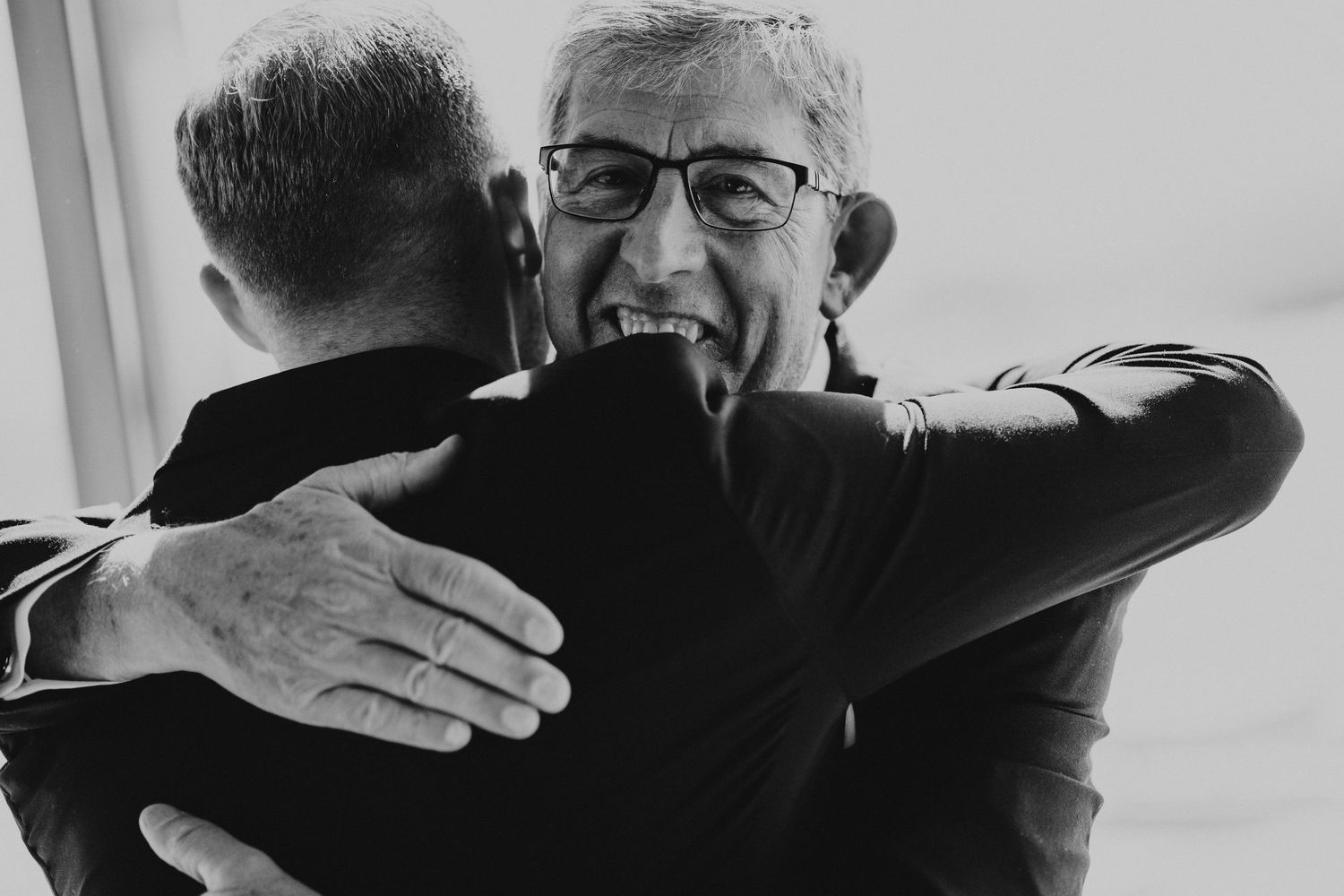 Emotional black and white photo of older man hugging younger man warmly at a wedding celebration.