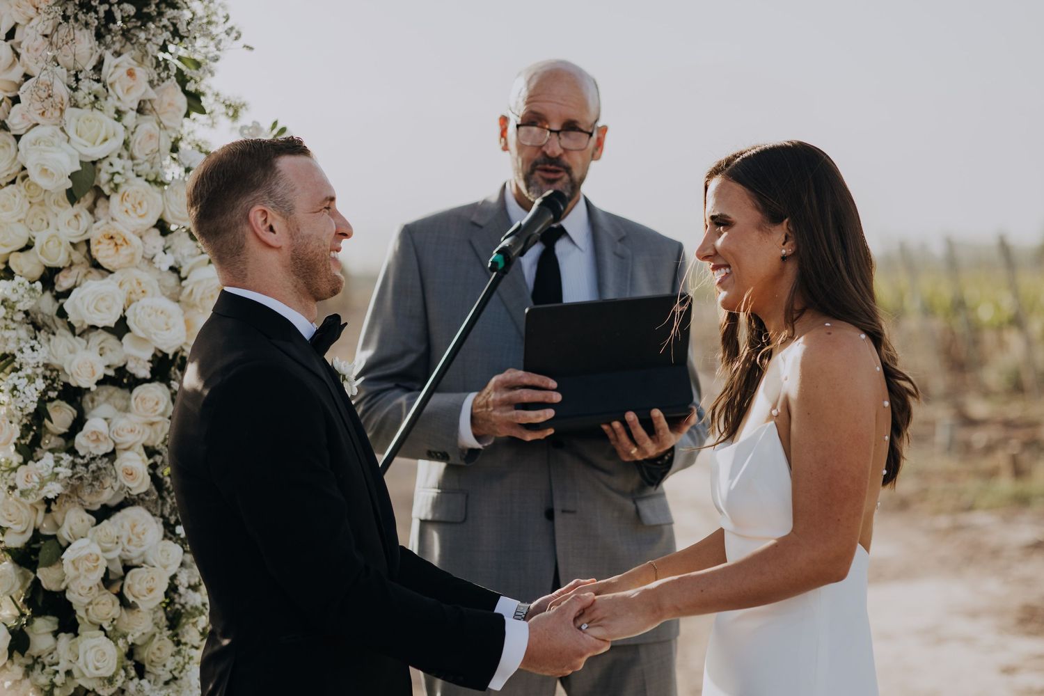 Couple exchanging vows at an outdoor wedding ceremony with a white floral arch and officiant.