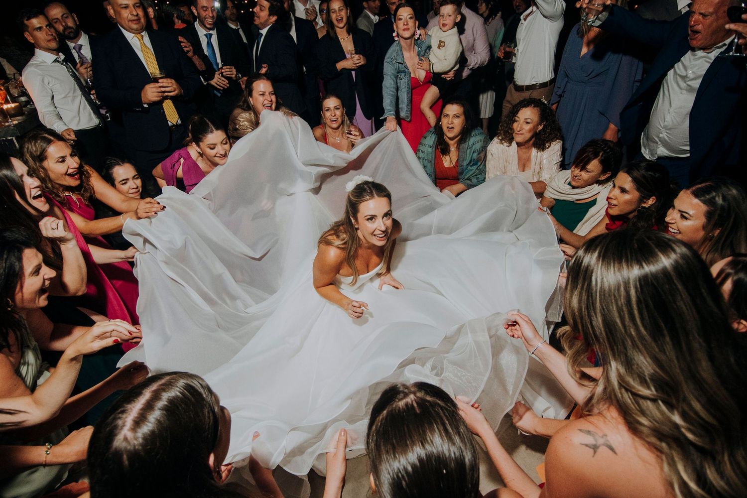 Aerial view of bride in white gown surrounded by guests lifting her dress at a lively wedding reception.