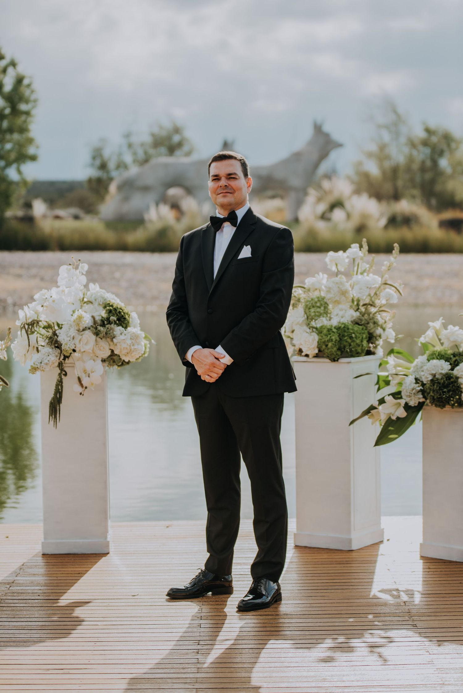 Groom in black tuxedo stands at outdoor waterfront wedding ceremony altar with white floral arrangements.