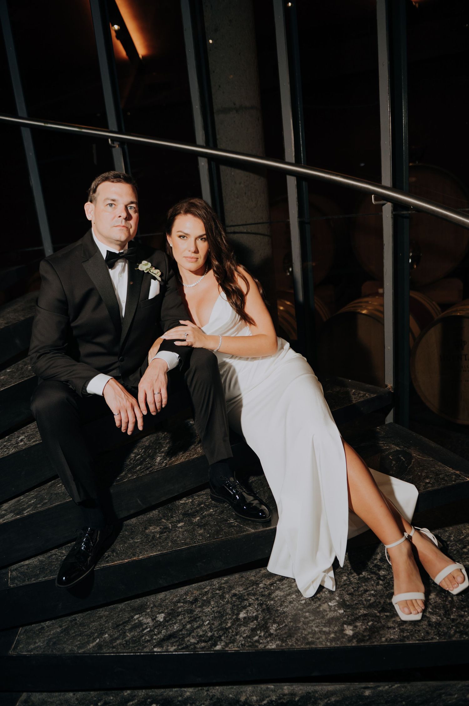 Bride and groom sit together on dark staircase at night in intimate wedding portrait.