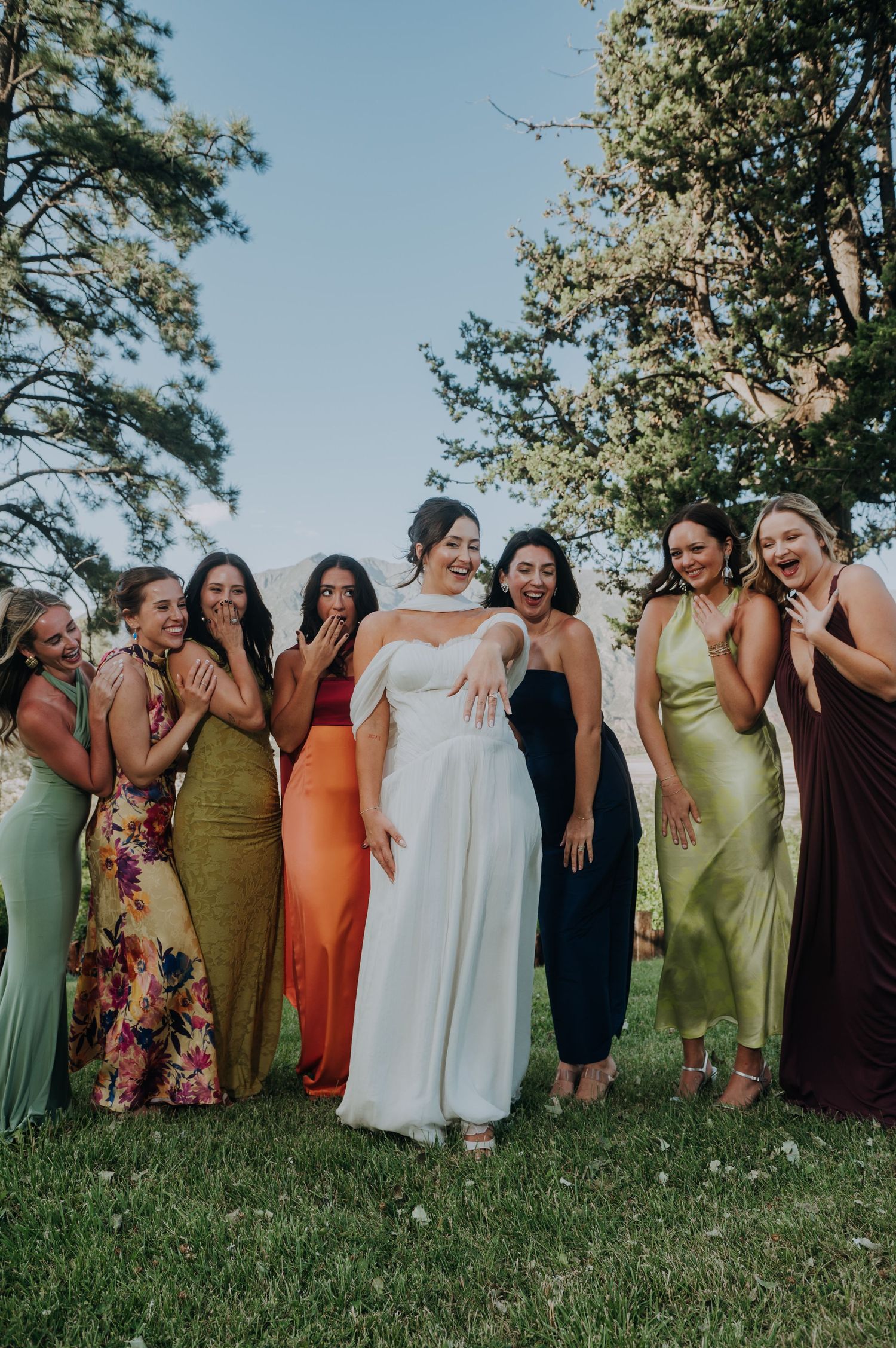 Bride in white gown laughs joyfully surrounded by bridesmaids in colorful mismatched dresses outdoors.