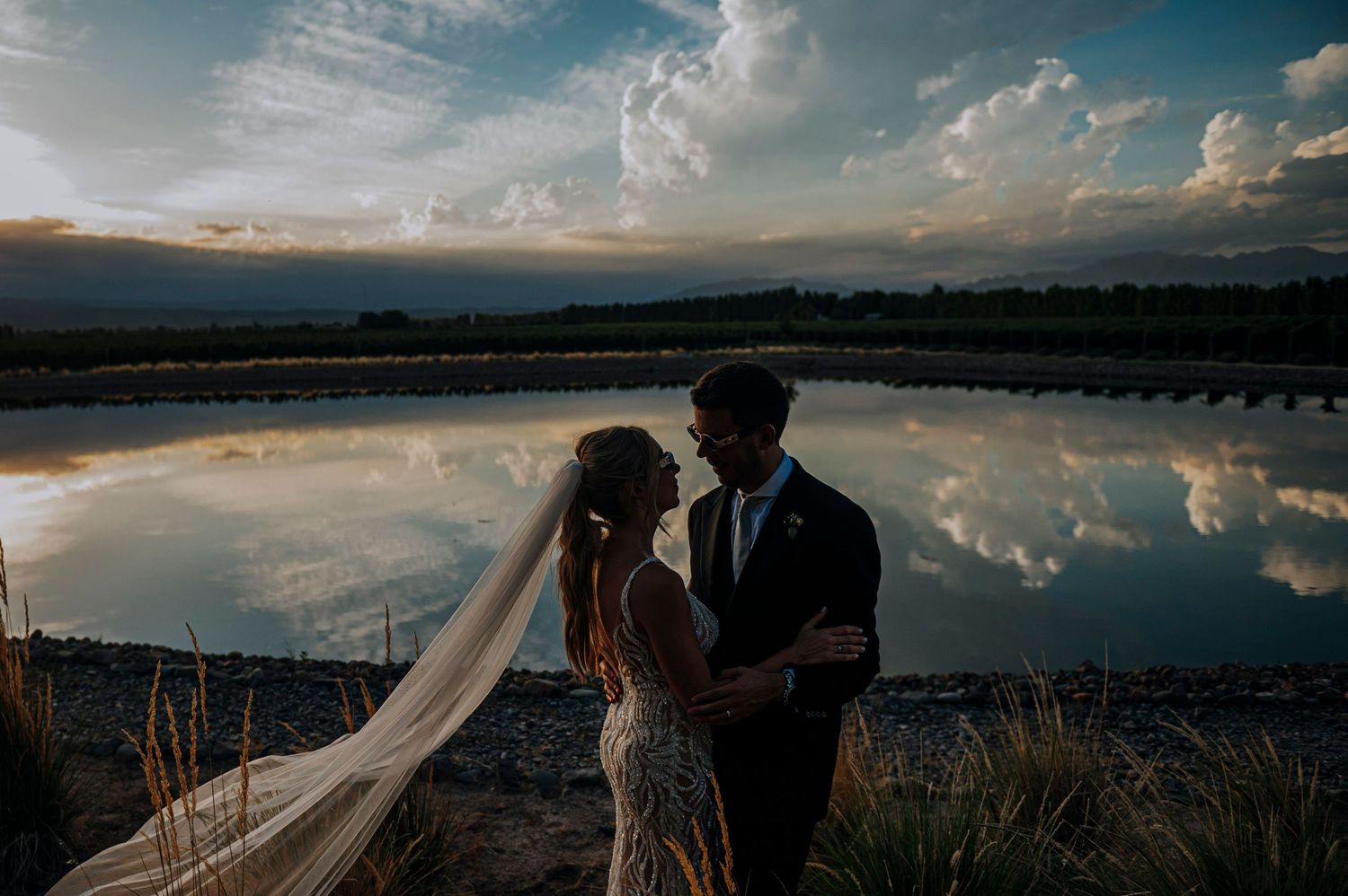 Bride and groom embrace at sunset beside a reflective pond with dramatic clouds mirrored in the water.