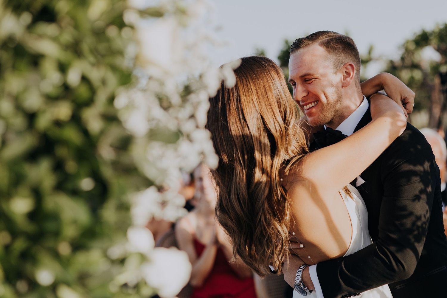 Smiling groom in tuxedo embracing bride outdoors during wedding celebration.