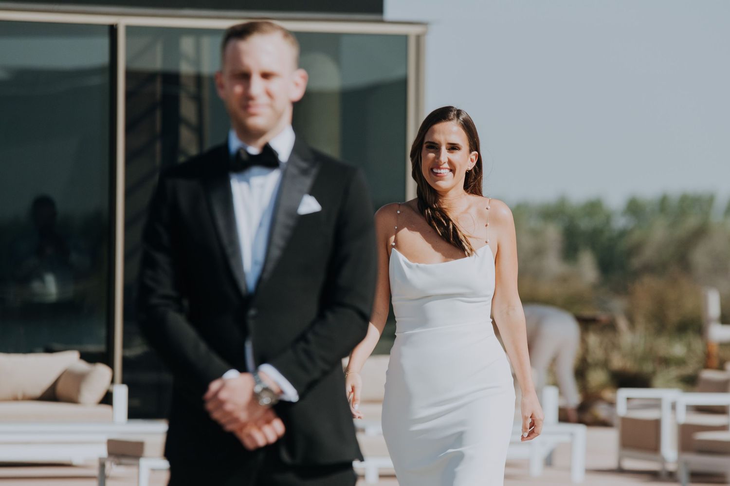 Bride in white gown approaching groom in tuxedo for a first look moment outdoors.