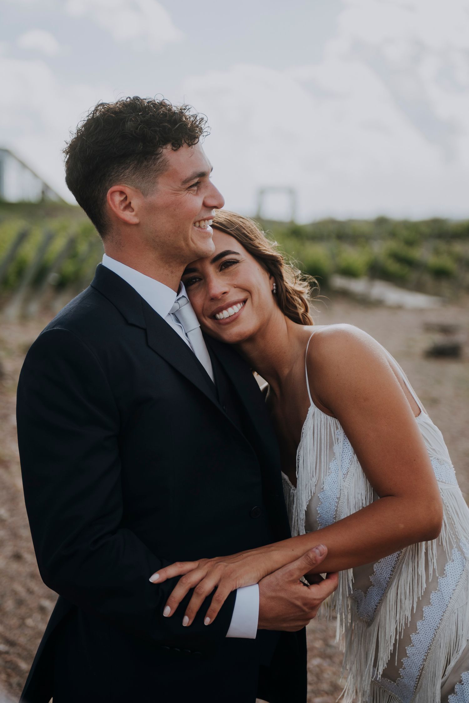 Smiling bride in fringe wedding dress leaning on groom in navy suit at vineyard wedding.