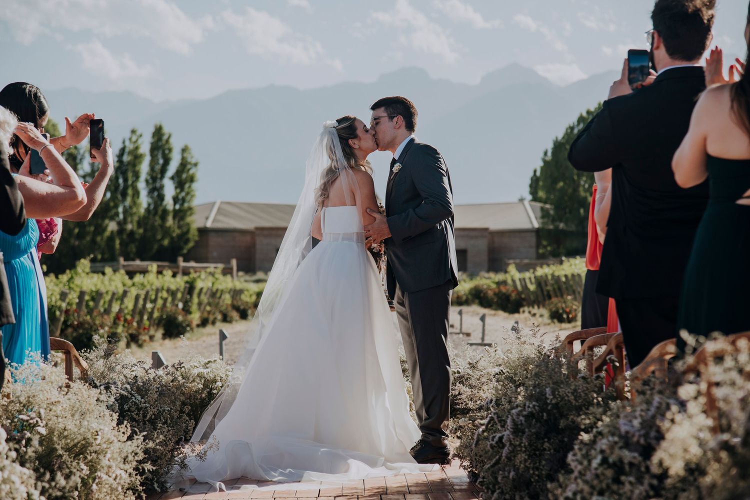 Bride and groom share first kiss at outdoor vineyard wedding ceremony with mountain backdrop and guests applauding.