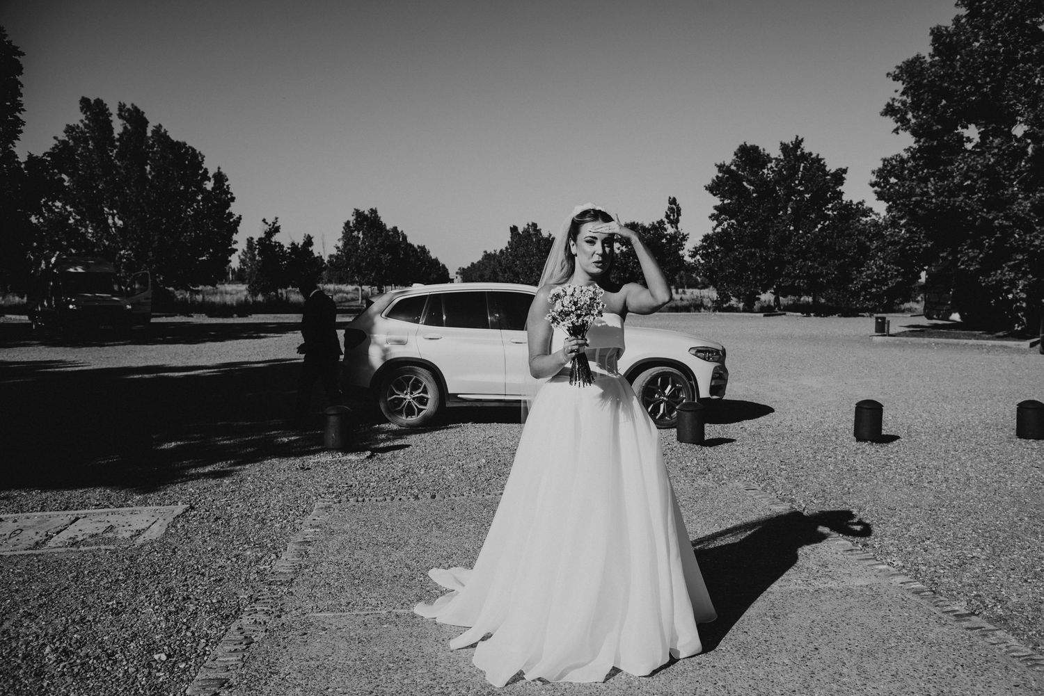 Black and white photo of bride in flowing gown standing in parking lot on wedding day.