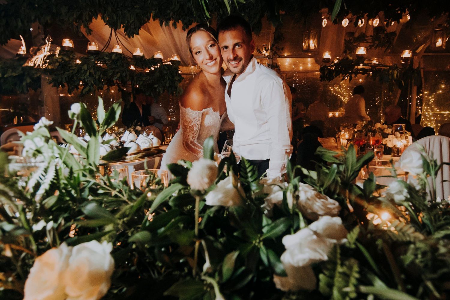Happy couple embraces behind lush white floral centerpieces at a warmly lit wedding reception.